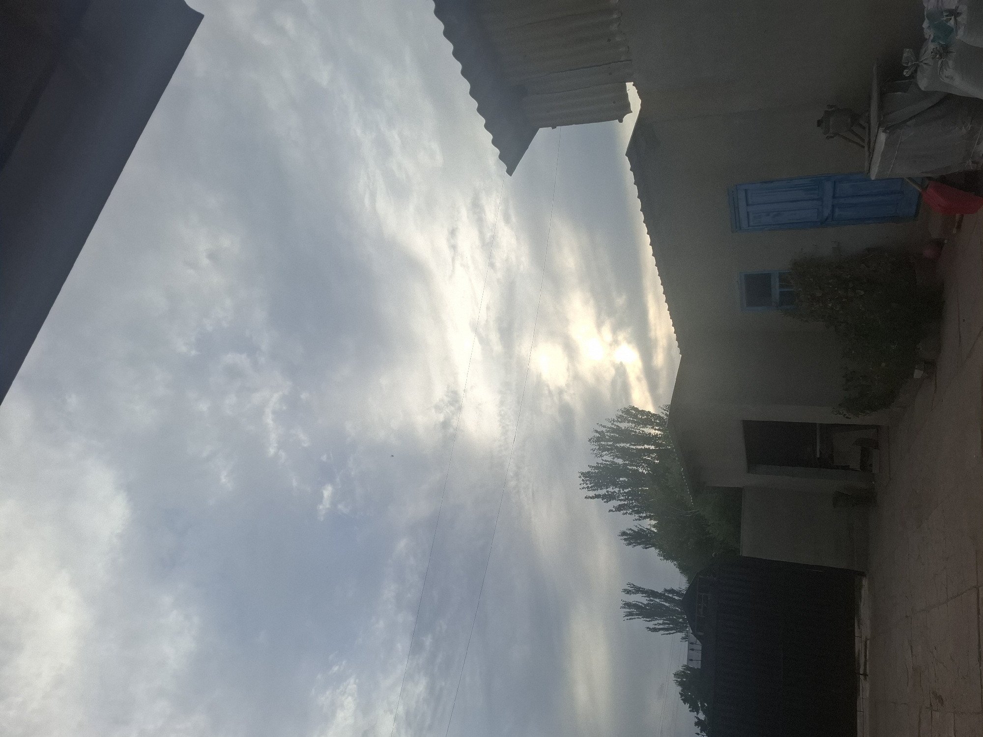 The image depicts a view of a residential area in Jeti-Ögüz, Kyrgyzstan, under a cloudy sky. The foreground shows a paved courtyard with a dark, corrugated metal fence on the left. To the right, a small, light-colored building with a corrugated roof is visible, featuring a blue wooden door and a small window. In front of this building, there is some greenery and scattered objects, including bags, a red shovel, and what appears to be a samovar or similar metal appliance on a table covered with a tarp. Behind the buildings, tall, slender trees are silhouetted against the sky. The sky itself is filled with a mix of grey and white clouds, with a bright, diffused light emanating from behind the clouds in the upper right quadrant, suggesting either sunrise or sunset. Power lines are visible stretching across the sky. The overall impression is that of a rural or semi-urban setting with simple architecture.