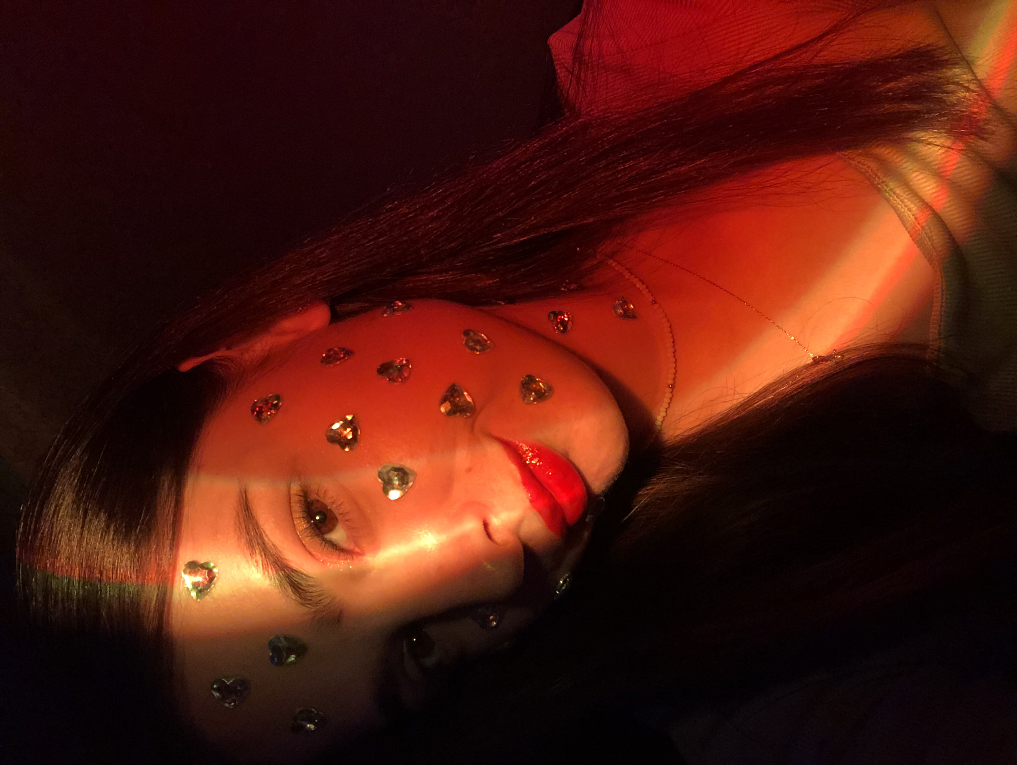 A close-up, high-angle, medium shot captures a young person's face adorned with heart-shaped rhinestones, set against a dark, red-hued background. The subject's left side is partially illuminated by a beam of light that refracts into rainbow colors across their forehead and cheek. They have dark hair, brown eyes, and are wearing bright red lipstick and a delicate necklace. The person is looking towards the camera with a slight smile. The scene appears to be indoors, with no specific identifiable location in Noviy, Russia, or any other city.