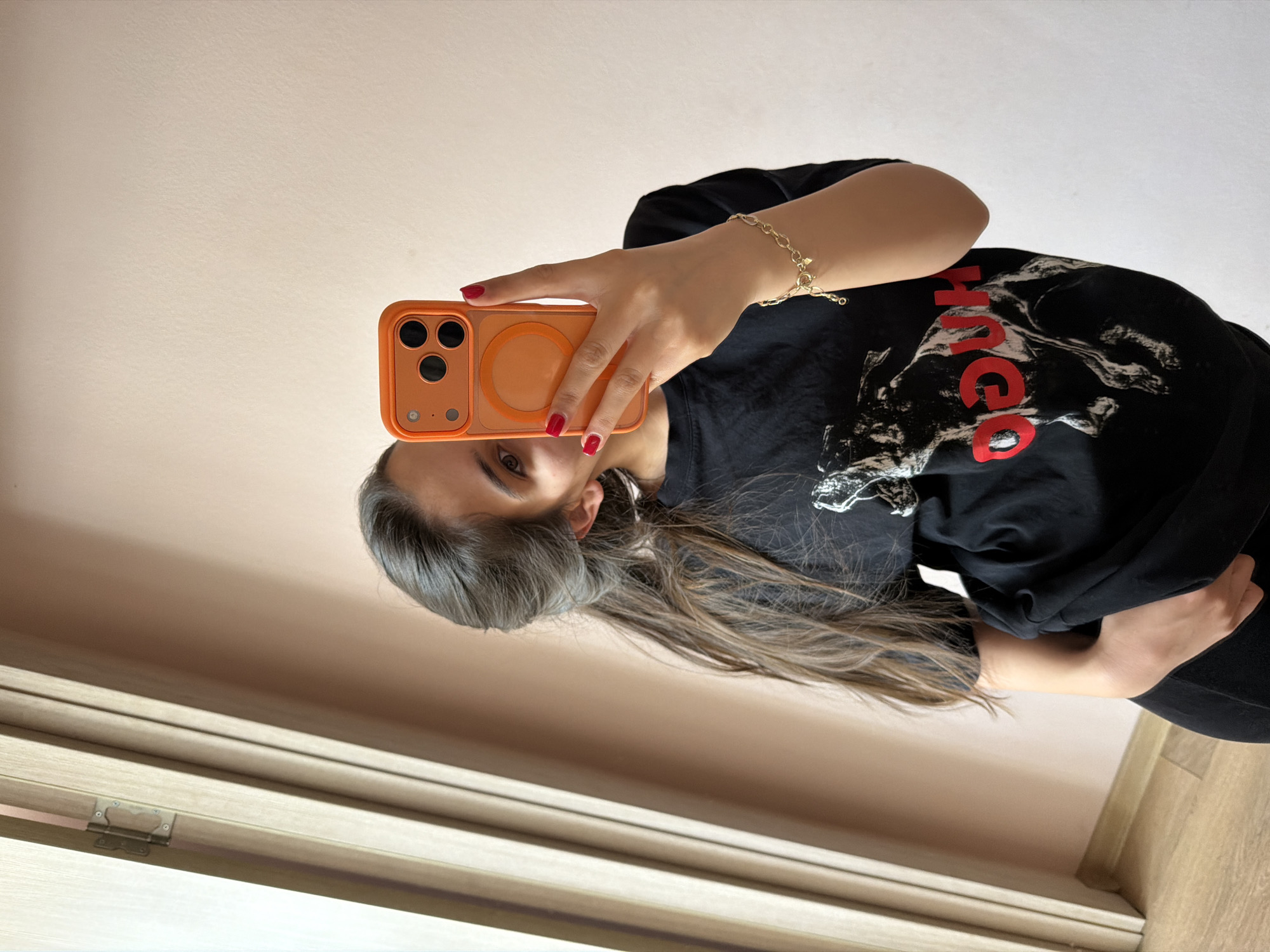 The image shows a woman indoors, holding an orange smartphone up to take a selfie. She is wearing a black t-shirt with a red and white design, and a gold bracelet on her left wrist. Her hair is long and brown. The background includes a window frame, a beige wall, and a wooden floor. There is no direct evidence to pinpoint the location in Rudartsi, Bulgaria; this determination would require further information or context.