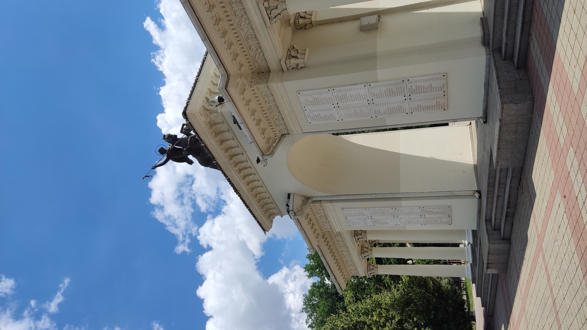 This media file features a close-up, low-angle shot of a building with classical architectural elements, set against a bright blue sky dotted with white, fluffy clouds. The building appears to be a memorial or a monument, possibly with a historical significance given the names inscribed on plaques mounted on its facade. The architecture includes a decorative frieze, columns, and an archway.
Prominently displayed on the building's exterior are several white plaques with rows of text, likely lists of names or historical records. These plaques are mounted on a light-colored wall. Above and to the left of the main structure, a dark bronze sculpture is visible, depicting a figure, possibly a warrior or a saint, on horseback, holding a spear or a lance. The sculpture has a halo, suggesting a religious or heroic figure.
The setting appears to be outdoors, under what seems to be a clear, sunny day. The lighting suggests it is daytime, likely late morning or afternoon, given the strong shadows cast by the architectural elements. The overall atmosphere conveyed is one of solemnity and remembrance, common for memorial sites. The background includes glimpses of lush green trees, indicating a park or a well-landscaped area. The ground in the foreground is paved with a checkered pattern of light and dark square tiles.
The orientation of the image is tilted, with the building and sky appearing at an angle. The location context provided is Krasnodar, Russia, which may help identify the specific monument depicted. No specific emotions are explicitly conveyed by the static image, but the subjects and setting evoke a sense of historical importance and reflection.
