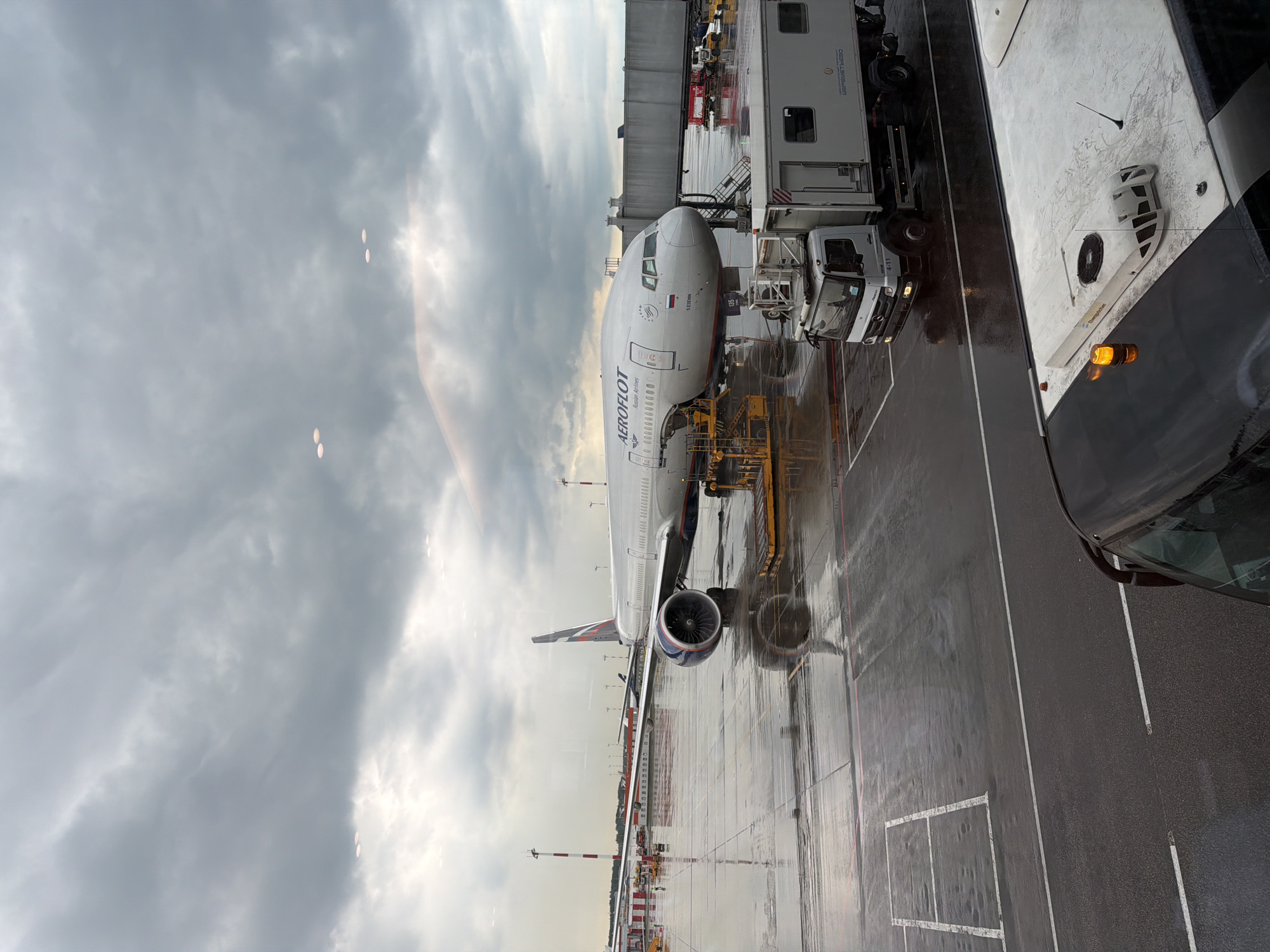 The image depicts an Aeroflot airplane parked at an airport gate, likely in Khimki, Russia. The scene is set on a wet tarmac, suggesting recent or ongoing rain, with the sky filled with dark, overcast clouds. The time of day appears to be late afternoon or early evening, given the low light.
The primary subject is a large passenger airplane, prominently displaying "Aeroflot Russian Airlines" on its fuselage. The aircraft is connected to a jet bridge, indicating boarding or disembarking activities. Several ground support vehicles, including a white truck and a baggage tug, are positioned near the aircraft, suggesting ongoing airport operations. A larger vehicle, possibly a bus or service van, is visible in the foreground, partially obscuring the view.
Notable details include the wet surface of the tarmac, reflecting the lights of the vehicles and the aircraft. Several circular, light-colored objects are visible in the sky, which could be lens flares or atmospheric phenomena. The weather appears gloomy and possibly cold.
The overall impression is one of a typical airport scene during inclement weather, with the focus on the parked aircraft and the surrounding operational equipment. No people are clearly visible in the image. The text "Aeroflot Russian Airlines" is the most prominent visible text. Another visible text on the jet bridge states "ШЕРЕМЕТЬЕВО", which is Sheremetyevo International Airport in Moscow, confirming the location.