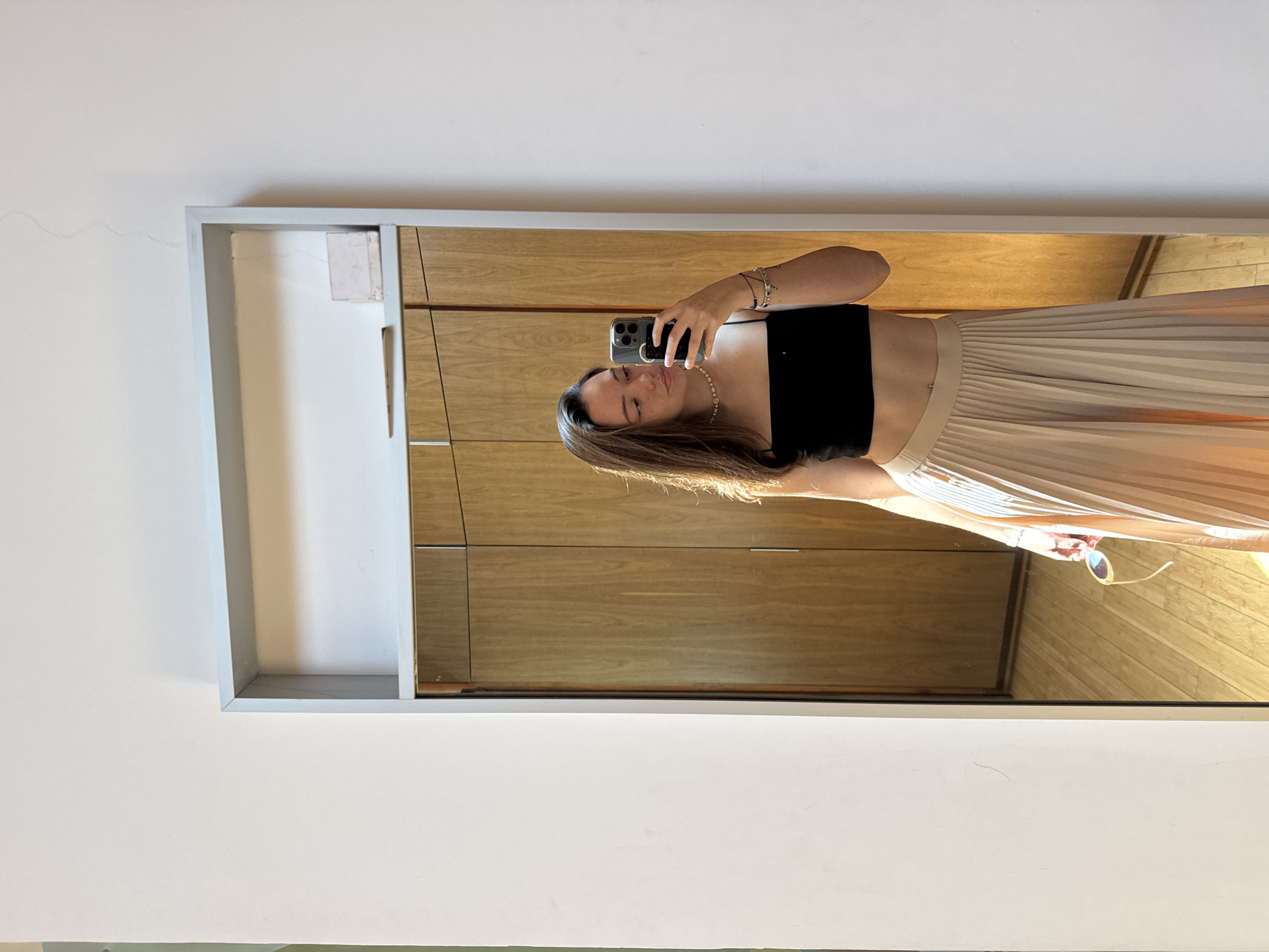 A person is taking a selfie in a mirror. The person is wearing a black top and a pleated skirt. The mirror is framed and reflects a wooden cabinet and a wooden floor. Sunglasses are also visible in the reflection. The location and city are not discernible from the image.