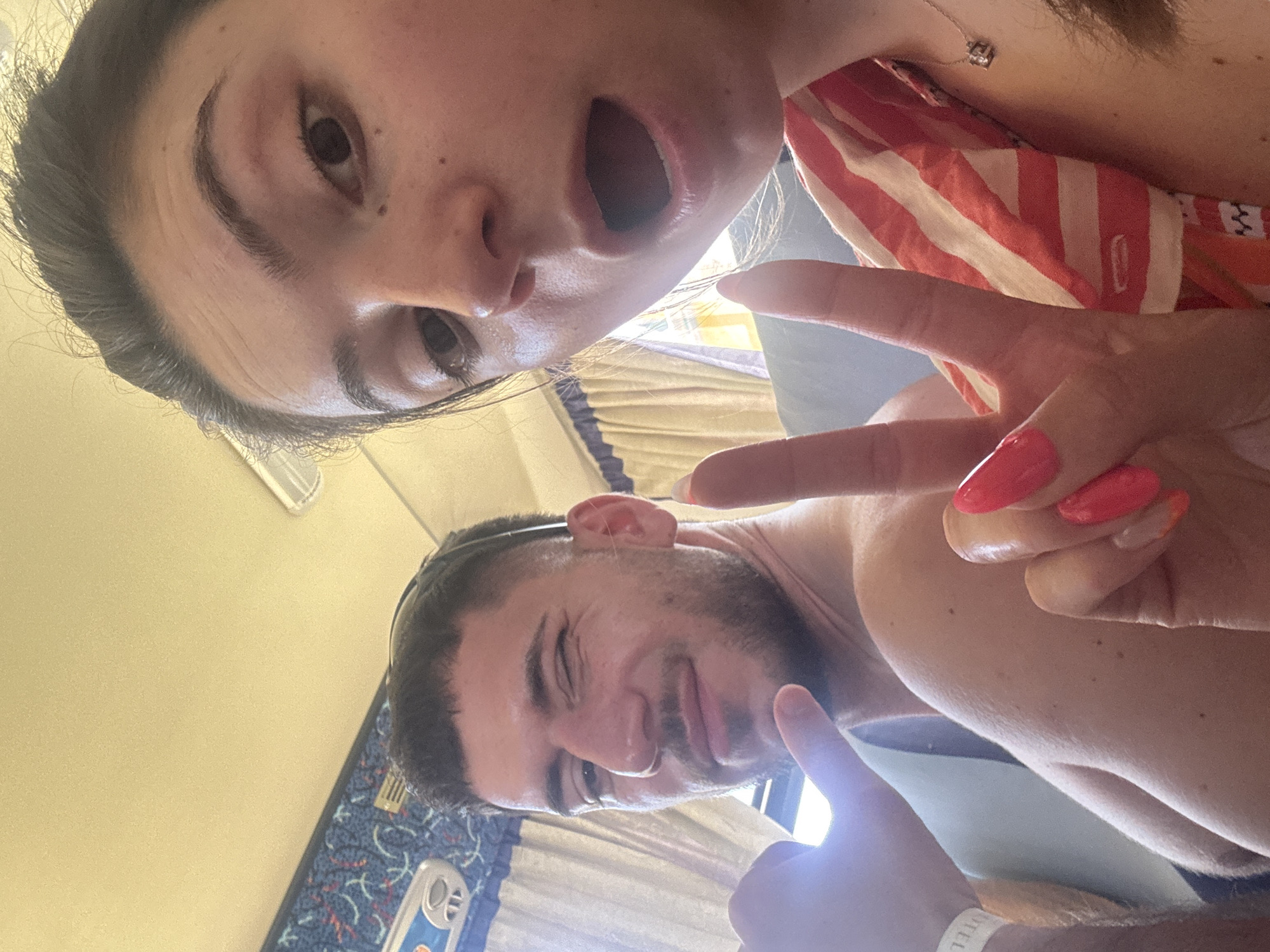 The image depicts two individuals, likely a couple, taking a selfie. The woman, positioned in the upper portion of the frame, has an open mouth and points with her index finger. She wears a necklace and a striped garment. Her fingernails are painted red. The man, in the lower portion of the frame, points with his index finger as well and wears a wristband labeled "HOTEL". The background shows what appears to be a curtained window and a bus or vehicle interior. The city and specific location are undetermined.