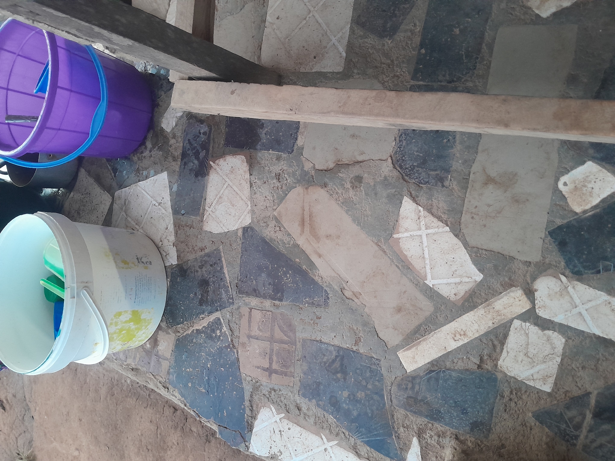 The scene is located in a utility area in Ifo, Nigeria, observed from an elevated perspective. The ground is paved with an irregular mosaic of broken ceramic tiles and stone fragments, primarily in shades of dark grey, light grey, and white, set within a light brown cementitious matrix; some white tile fragments display etched or painted cross-hatch patterns. In the upper left, a white plastic bucket, exhibiting yellow-green stains and some legible text including "PT Maligana," contains a blue plastic scoop, a green sponge, and a clear-green plastic item. To its right, a purple plastic bucket with a light blue handle rests on the tiled surface, holding a blue plastic tool and a thin, dark rod; a small amount of liquid is visible on the ground around its base. Partially obscured metallic pots are present in the background between the two buckets. A light-colored wooden beam extends vertically along the right side of the frame. No individuals are present.