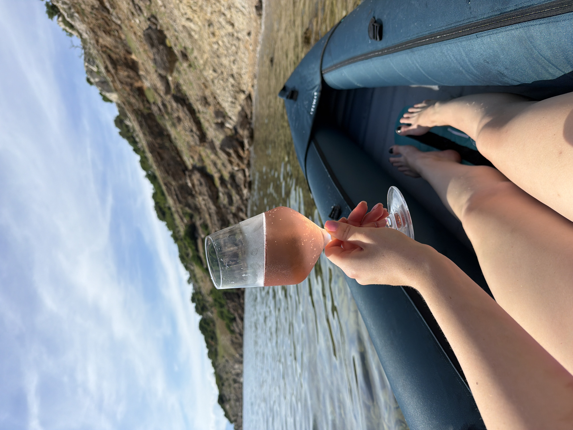 In an over-the-shoulder shot, a person's arm holds a stemmed wine glass filled with rosé wine, angled towards a rocky, tree-covered coastline under a partially cloudy sky. The person is seated in an inflatable kayak, with their legs visible in the foreground. The water is calm and clear, with sunlight reflecting off its surface. The kayak is a dark blue, and its interior is also visible. The scene appears to be in a natural, coastal environment.