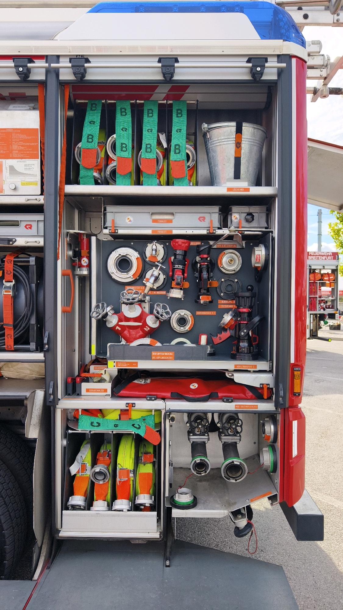 This is a detailed, eye-level, full shot of the side storage compartment of a red and white fire truck. The compartment is open, revealing organized equipment. In the top section, four coiled green hoses with orange connectors are hung vertically from metal clasps. To the right of the hoses is a silver metal bucket. Below this section, a large grey panel is filled with various fire hose fittings, nozzles, valves, and other connectors, predominantly red and silver with orange accents. In the bottom section, to the left, are more coiled green hoses with orange connectors. To the right, two large black hoses with green and silver connectors extend downwards from the truck. The truck appears to be parked outdoors on a paved surface, possibly a street or parking lot, with a hint of green foliage and sky visible in the background. The truck has "FEUERWEHR BREITENFURT" written in white lettering on a red background on its side.