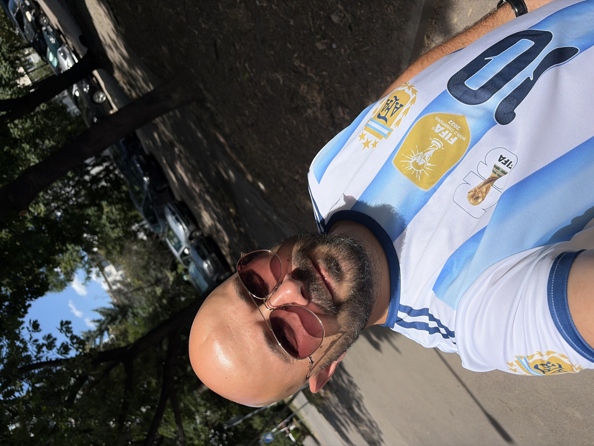 A man with a bald head and a beard is taking a selfie outdoors. He is wearing an Argentina national football team jersey with the number 10 on it, and the FIFA World Cup 2022 champions badge. He is also wearing aviator sunglasses with pinkish lenses. The man is standing on a sidewalk next to a road with parked cars. Trees are visible in the background, as well as buildings and a blue sky. The lighting suggests it is daytime and sunny. The scene appears to be in a residential or urban area.
