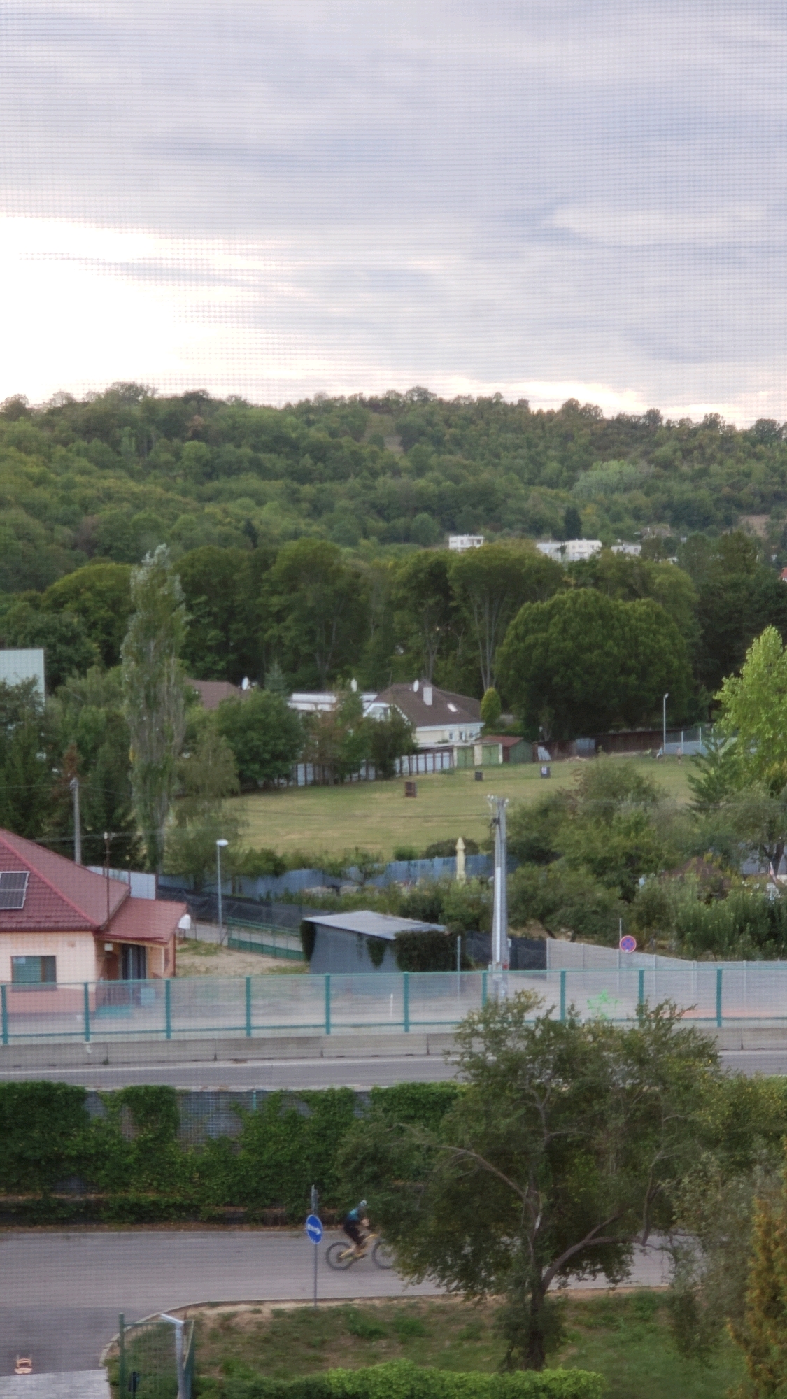This is a photograph of a residential area in Prešov, Slovakia, likely taken from an elevated position, possibly a window. In the foreground, a paved road is visible with a person on a bicycle riding on it. A large tree with green foliage partially obscures the view of the road and cyclist. A street sign with a blue circle and a white arrow is positioned near the road. Further into the scene, a row of green hedges lines a road or pathway. Beyond the hedges, a light-colored fence separates an area with grass and some smaller buildings from a road. In the middle ground, several houses with red and light-colored roofs are nestled among trees. One house, in particular, has a solar panel on its roof. Further in the background, a densely forested hill rises, with the sky visible above. The sky is overcast with soft clouds. The overall impression is of a quiet, suburban environment with a blend of residential buildings and natural greenery.
