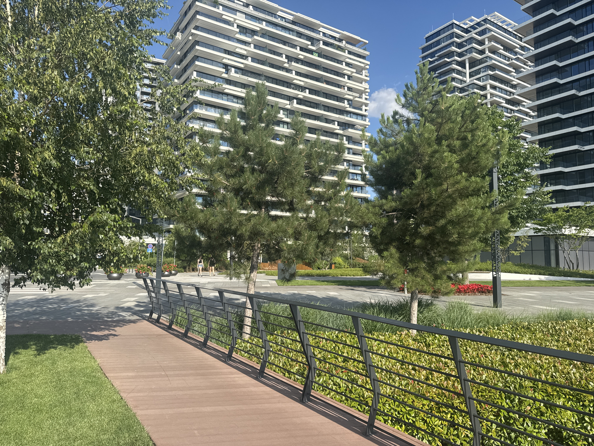 The image captures a sunlit outdoor scene in Belgrade, Serbia, featuring modern architecture and landscaping. In the foreground, a wooden boardwalk with a dark metal railing runs diagonally across the frame, bordered by lush green grass on the left and dense green shrubs on the right. Several trees, including a birch with a white trunk and a pine tree, are scattered throughout the scene, casting dappled shadows on the boardwalk and the paved area beyond.
Behind the greenery, a paved walkway or plaza is visible, with a few people casually walking. The architecture is characterized by tall, modern buildings with repeating horizontal lines of balconies and windows, giving them a stacked appearance. The buildings are predominantly white and glass, reflecting the bright daylight. The sky is a clear blue with a few white clouds, suggesting pleasant weather.
In the mid-ground, several decorative planters with red flowers add pops of color to the urban landscape. A modern-style lamppost with a geometric design stands near the planters. The overall impression is one of a well-maintained urban public space, possibly a park or a residential complex with integrated green areas and contemporary design. The time of day appears to be mid-morning or afternoon, given the bright sunlight and distinct shadows. No visible text is present in the image.