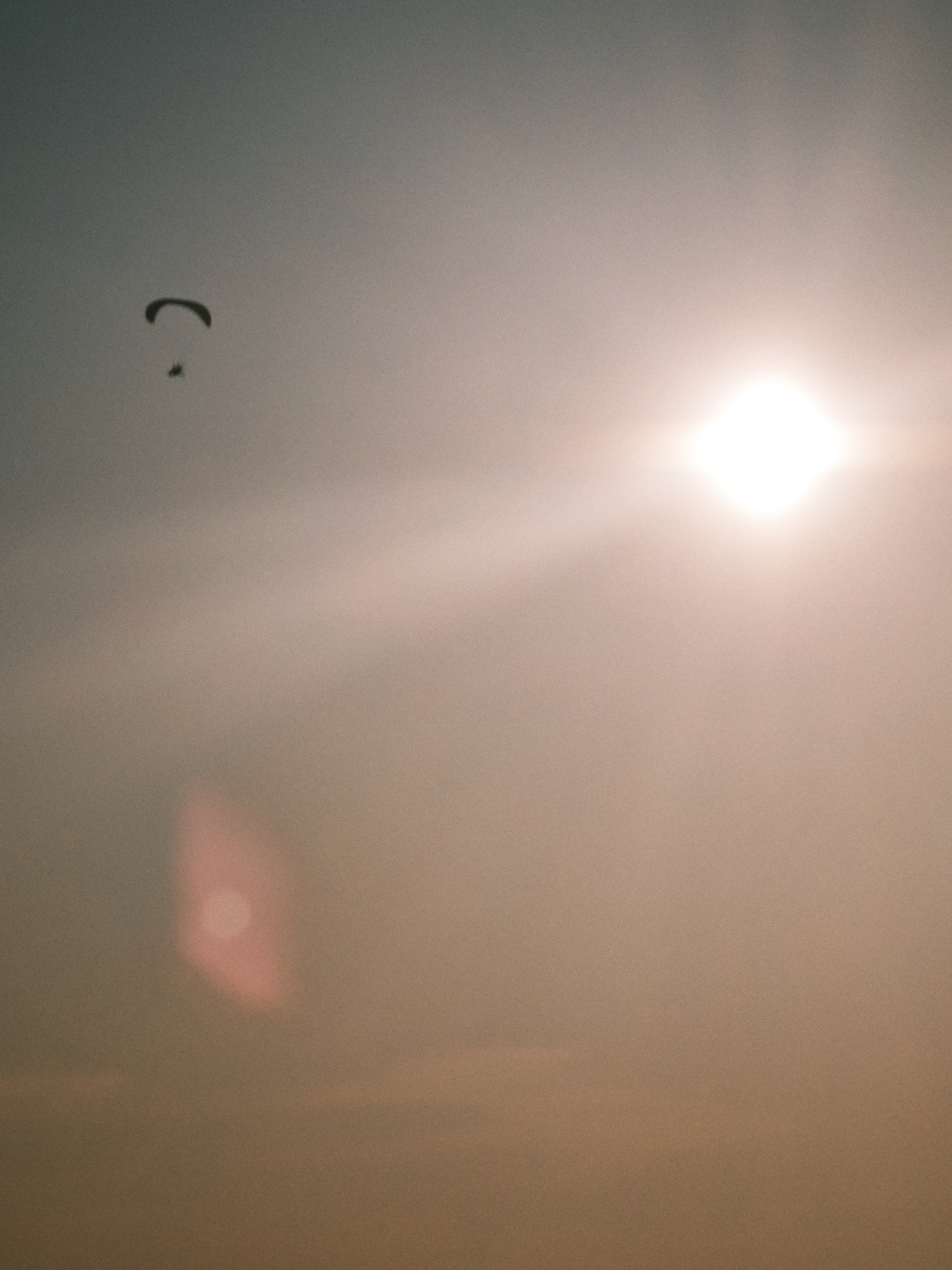 The image depicts a paraglider in silhouette against a hazy sky, with the sun prominently visible as a bright, square-shaped lens flare. The paraglider, a small dark shape, is positioned in the upper left quadrant of the image, with its canopy curved and a smaller figure visible beneath it. The sky is a gradient of soft colors, transitioning from a muted grey-blue at the top to a warm, peachy tone towards the bottom, likely indicating sunset or sunrise. A large, bright sun, emitting rays of light, is located in the upper right portion of the frame. A reddish, out-of-focus light flare is visible in the lower left quadrant. The overall atmosphere is hazy and somewhat dreamlike due to the atmospheric conditions and the lens flare. There is no indication of the city of Lesosibirsk, Russia, in the image.