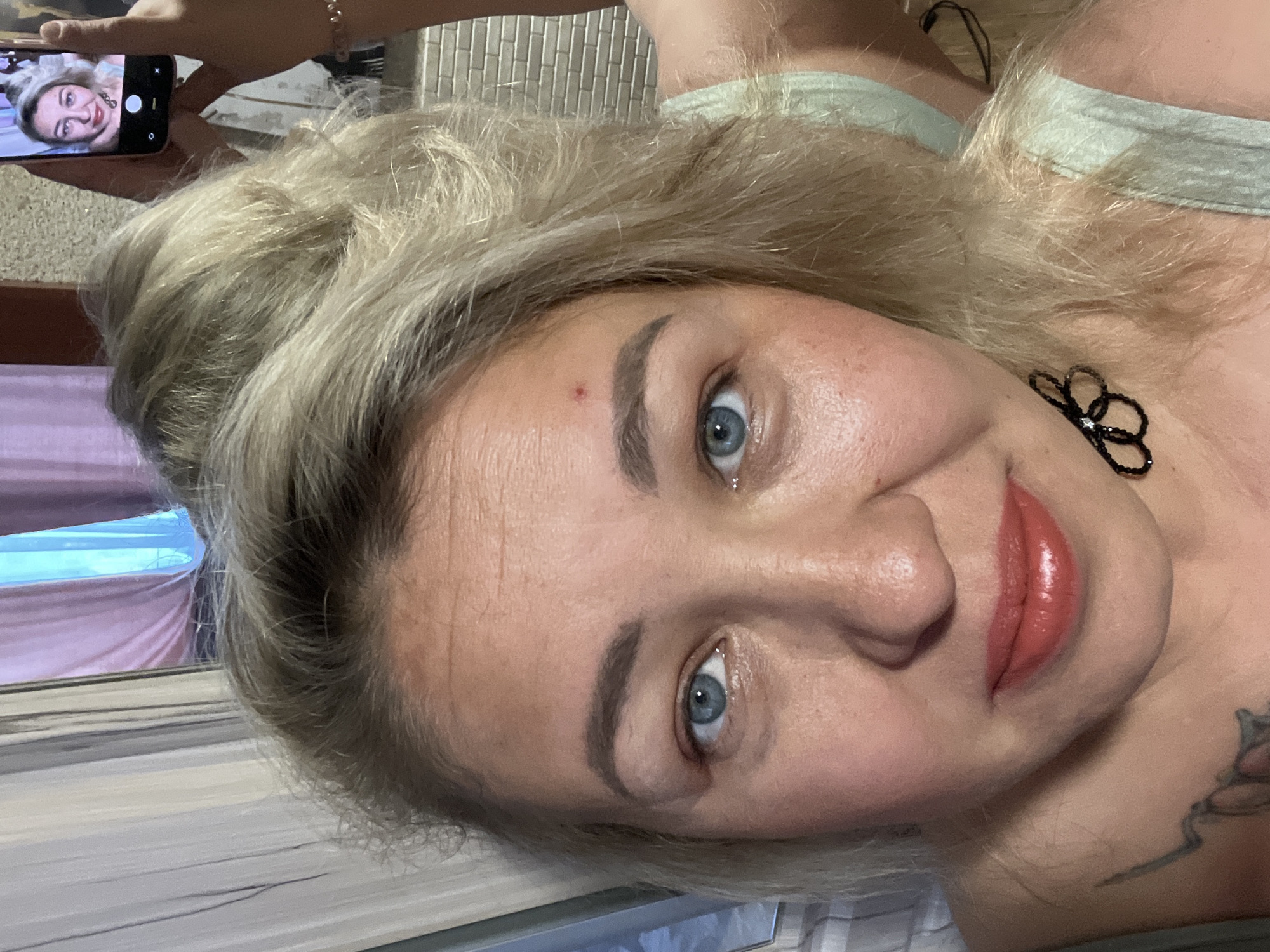 A woman with blonde hair and blue eyes is posing for a selfie. She is holding a phone in her left hand, which displays a smaller image of herself. She has a red mark on her forehead and is wearing black flower-shaped earrings. A glimpse of a tattoo is visible on her upper chest. The background is blurred, revealing a window with curtains. The scene appears to be indoors. There is no discernible information to confirm that the picture was taken in the city of Chaikovsky, Russia.
