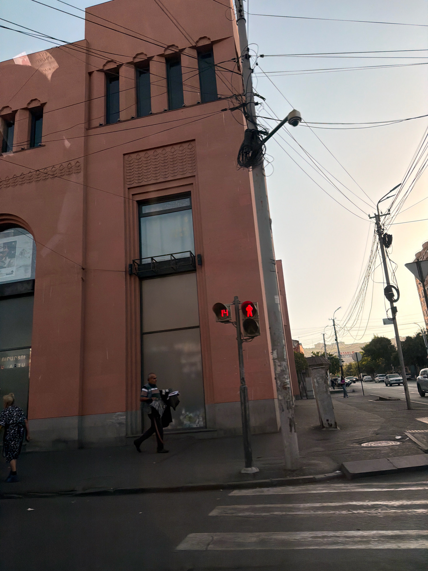 The image captures a street scene in Yerevan, Armenia, at what appears to be either dawn or dusk, given the soft lighting. A distinct salmon-pink building dominates the left side of the frame, featuring a series of recessed windows and decorative architectural elements. In front of this building, a pedestrian crossing is visible on the asphalt road.
On the sidewalk, a man is walking away from the viewer, carrying a bundle of dark clothing over his arm. He is wearing a horizontally striped shirt and dark pants. Further to the left, a woman in a patterned dress is walking towards the viewer.
A traffic light with red illuminated figures, indicating "don't walk" for pedestrians, stands prominently in the center of the image. Above the traffic light, a security camera is mounted on a pole amidst a complex network of overhead wires, characteristic of urban infrastructure.
The background reveals more of the city, with other buildings and trees lining the street. Several cars are visible on the road, suggesting ongoing traffic. The sky is a pale, hazy blue, transitioning to a lighter tone towards the horizon. The overall impression is of an ordinary urban moment unfolding in a specific Armenian locale.