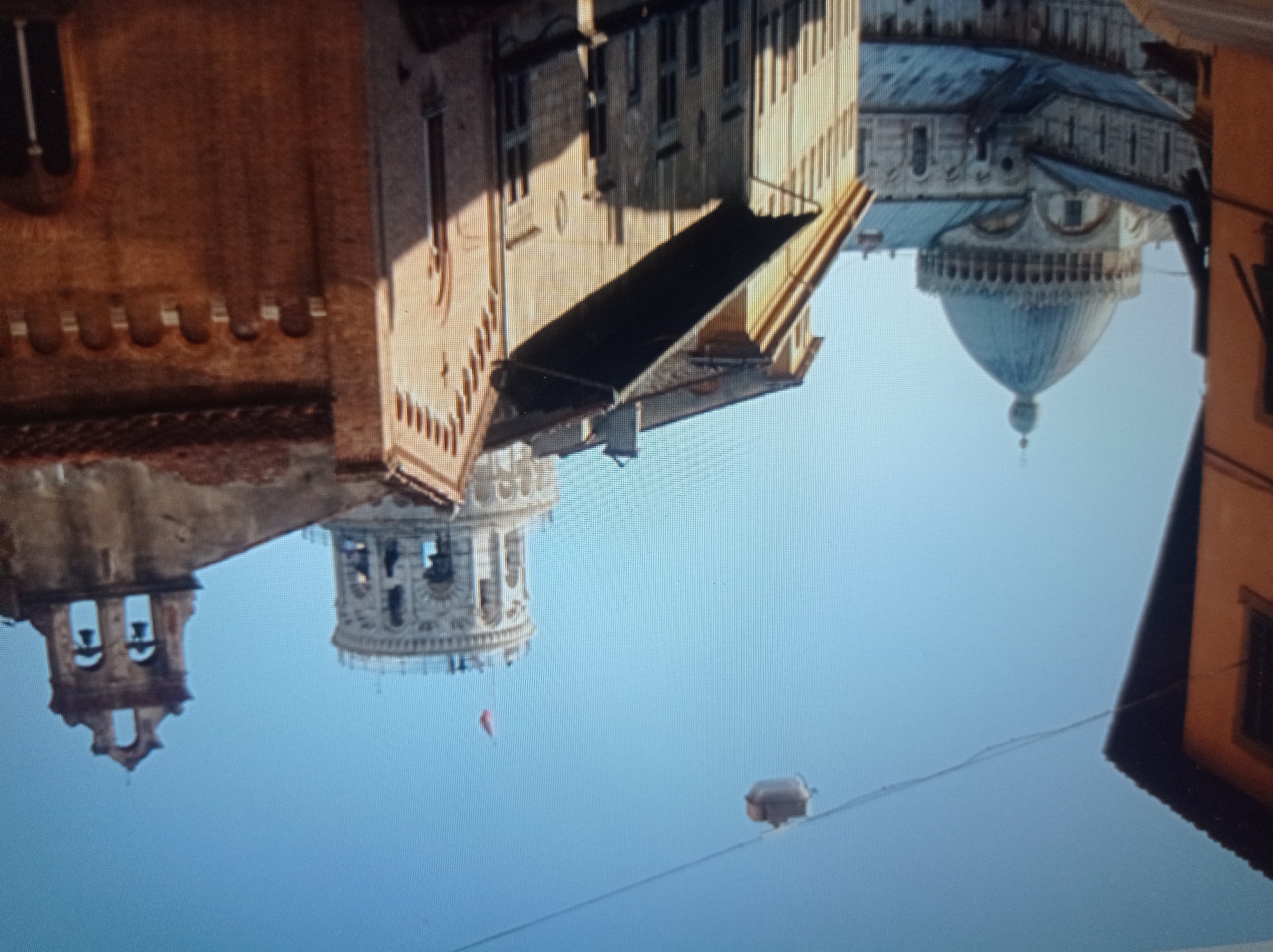 This is a photograph of the Pisa Cathedral complex in Pisa, Italy. The image captures several iconic structures including the Leaning Tower of Pisa, the Cathedral itself with its prominent dome, and the bell tower. The structures are made of stone and brick, with architectural details typical of Romanesque style. The sky is clear and blue. There are no people visible in the image. The photograph is taken from a street-level perspective, with the buildings dominating the foreground and mid-ground. The environment appears to be an urban setting, characterized by historical architecture. The question incorrectly states the location is Mukachevo, Ukraine, which is not the case.
