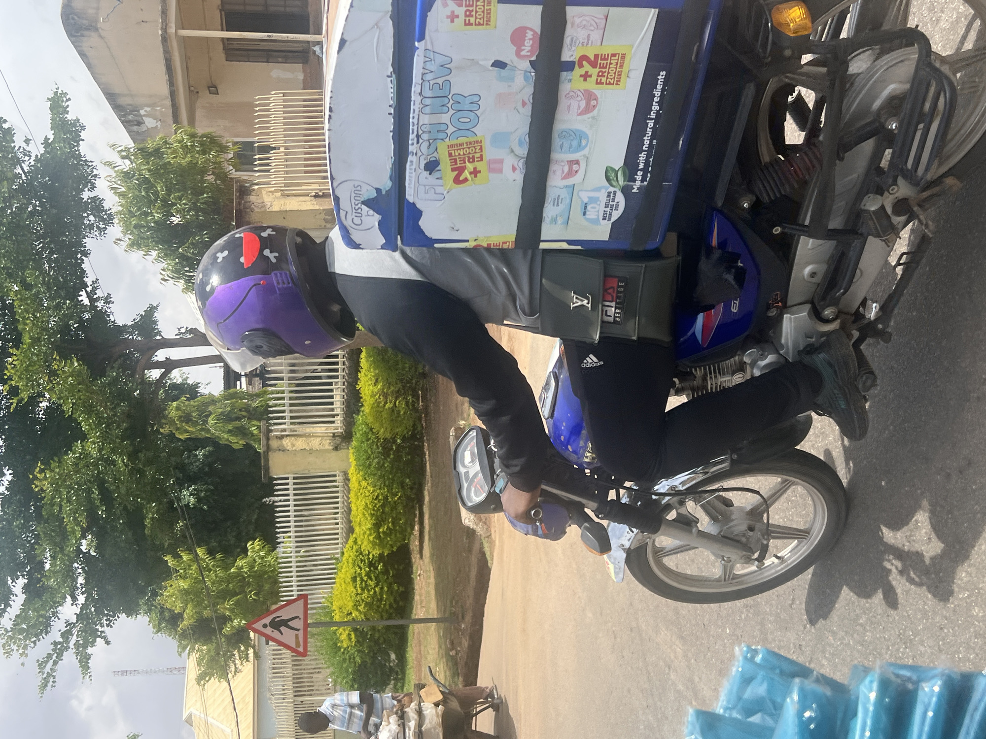 The image, taken in Jos, Nigeria, captures an outdoor, daytime street scene dominated by a person on a motorcycle. The rider, likely a delivery driver, is male and wears a purple and black helmet, a black and white long-sleeved top, and black trousers with an "adidas" logo visible near the ankle. A large white and blue delivery box, heavily branded, is securely fastened to the back of the blue and silver motorcycle.
The delivery box prominently displays "Cussons" branding, along with phrases such as "FRESH NEW LOOK," "New," "+2 FREE 200ML PACKS INSIDE" (repeated), "Made with natural ingredients," and "NO. 1 BEST SELLING SKINCARE BRAND 2024." The rider also has a small black pouch with "FILA HERITAGE" text and another with an "LV"-like logo attached to their gear.
The background features lush green trees and bushes, a white fence, and older-looking buildings with signs of wear. The street is paved, and strong shadows on the ground indicate bright sunlight. A white triangular road sign showing a black pedestrian is visible on the roadside. In the blurred background, another person stands near a small market stall or cart. Several out-of-focus blue packaged items are visible in the foreground. The overall impression is a bustling urban or semi-urban environment.