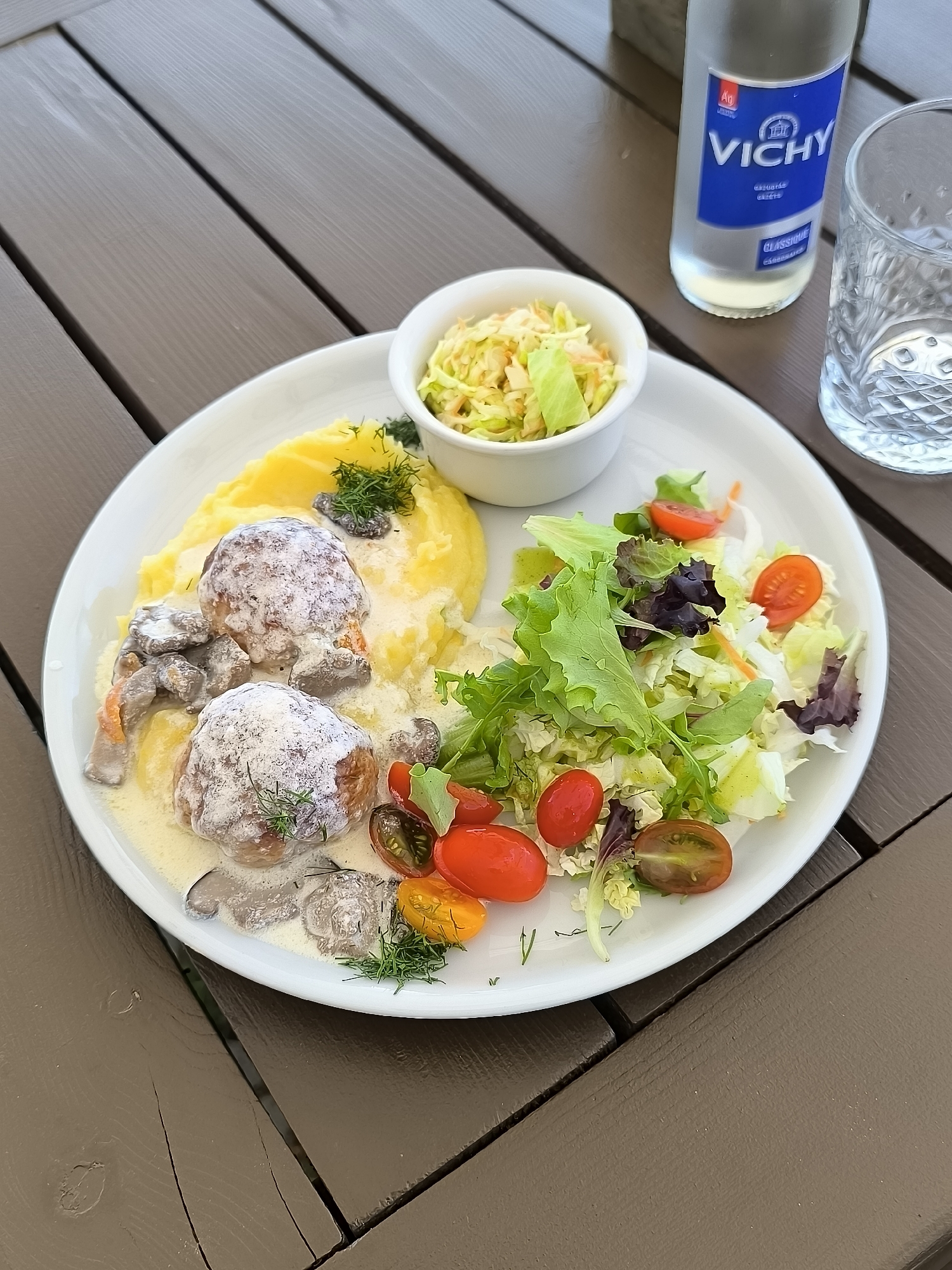 On a brown wooden table, a white plate holds a meal. The plate contains two meatballs covered in a creamy mushroom sauce, a generous portion of mashed potatoes, and a side salad with cherry tomatoes and mixed greens. A small white bowl of coleslaw sits at the top of the plate. To the right of the plate are a glass of water and a bottle of Vichy water. The setting appears to be outdoors, possibly a restaurant in Neringa, Lithuania, given the Vichy water bottle, a popular brand in the region, and the wooden table's style, reminiscent of furniture often found in outdoor dining areas in seaside locations.
