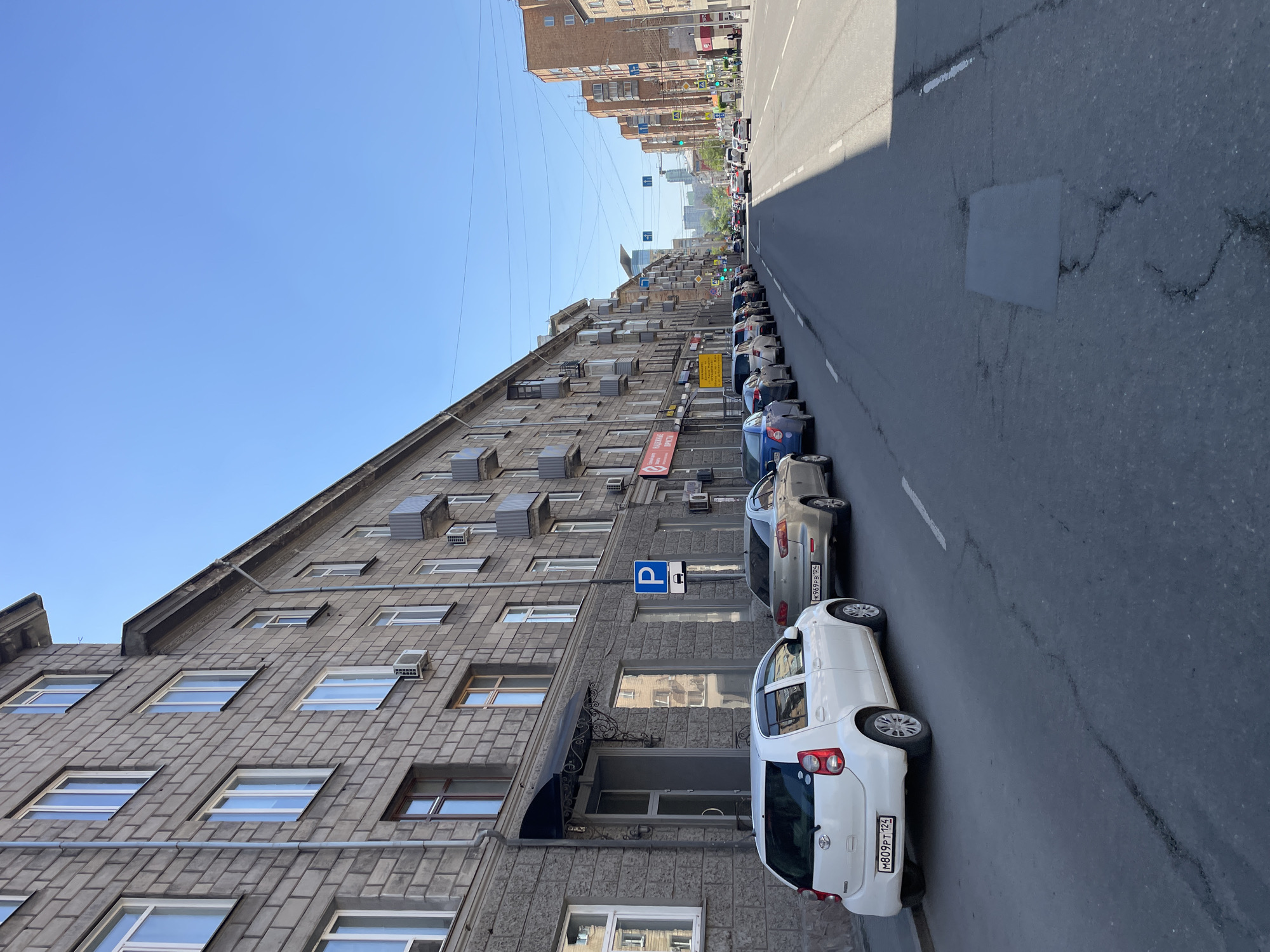 The image captures an urban street scene in Krasnoyarsk, Russia, under a clear blue sky. The perspective is looking down a long, straight street lined with buildings.
On the left, a multi-story building dominates the foreground, constructed of gray, textured stone or concrete blocks. It features numerous rectangular windows, many with white frames, and several external air conditioning units mounted on its facade. Below the building, cars are parked parallel to the curb.
The street itself is wide and made of asphalt, showing signs of wear with visible cracks and patches, including a distinct rectangular patch in the foreground. White lane markings are visible on the road surface.
A line of parked cars extends into the distance along the left side of the street. In the immediate foreground, a white car is parked, its rear visible with a Russian license plate "M809PT 124". Behind it, a silver car is parked, displaying the license plate "H969PB 124".
Notable details include:
* **Time of Day/Weather:** Bright daylight with clear blue skies, suggested by the strong shadows cast by the building across the street.
* **Signs:** A blue parking sign with a white 'P' and a rectangular symbol is visible on the sidewalk next to the parked cars. Other street signs and traffic lights are visible further down the street, including blue signs with white symbols. A red sign with a white graphic is on the building, and a yellow sign with unreadable text is also present.
* **Architecture:** Beyond the immediate gray building, taller brick-faced buildings of varying styles are visible further down the street, contributing to the urban setting.
* **Infrastructure:** Overhead power lines crisscross the sky above the street, indicative of an urban environment.
The scene depicts a typical busy city street, likely during the day, with parked vehicles and urban infrastructure. No people are clearly visible.