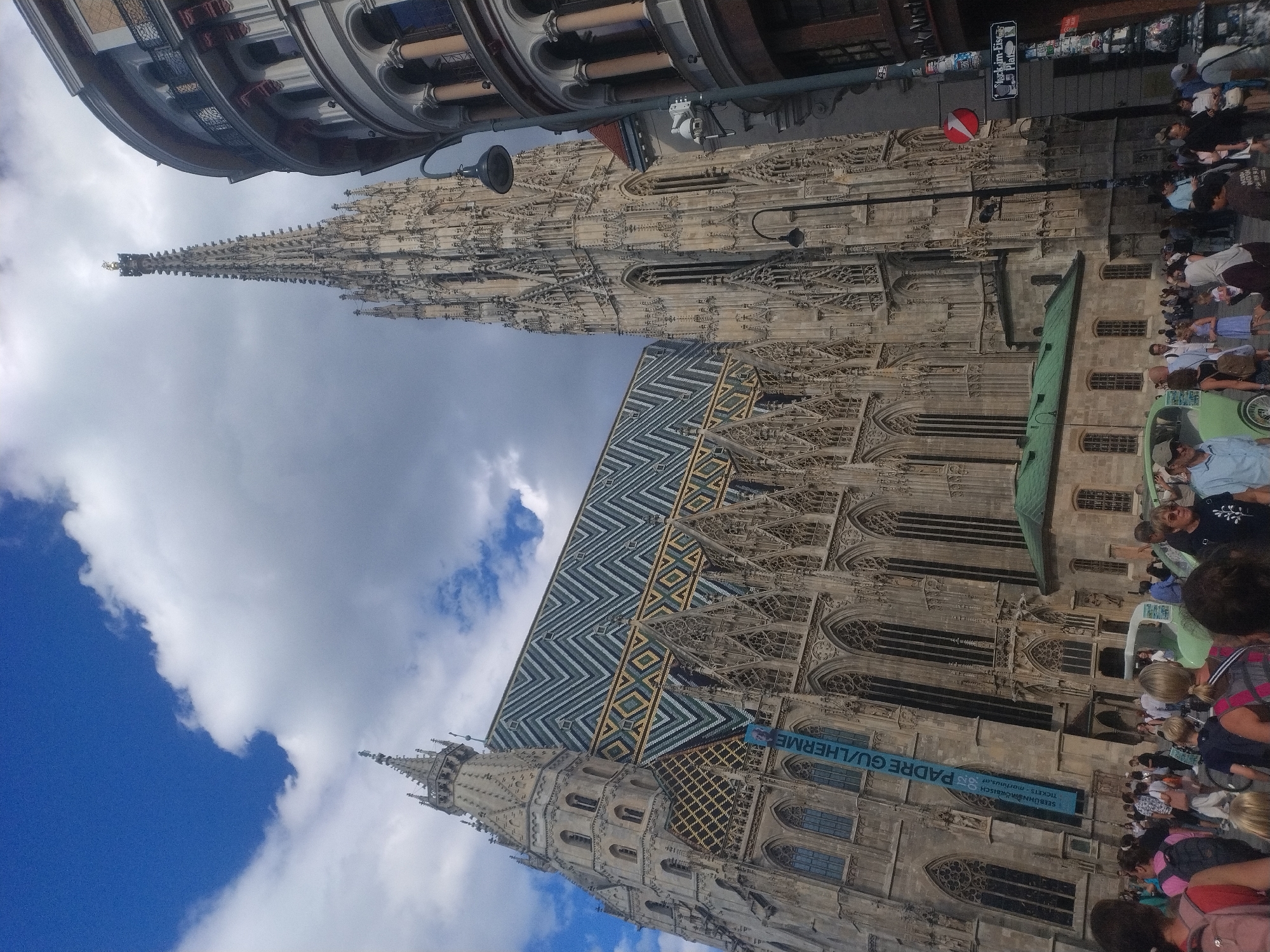 This is a wide-angle, ground-level shot of St. Stephen's Cathedral in Vienna, Austria, under a cloudy blue sky. The cathedral's Gothic architecture is prominently featured, with its tall, ornate spire dominating the right side of the frame, and the elaborately tiled roof of the nave visible on the left. A large, blue banner with white lettering is draped down the side of the cathedral. In the foreground, a crowd of people are milling about, some dressed in casual clothing, others in more formal attire. Several individuals are looking towards the camera, while others are engaged in conversation or observing the cathedral. A light green vintage car is parked in the middle ground, to the left of the crowd. To the right of the cathedral, a modern building with arched windows and balconies is visible. A street lamp stands in the foreground, to the right of the vintage car. The street is paved with cobblestones, and a street sign with German text is visible on a pole to the far right. The overall scene depicts a bustling urban environment with a significant historical landmark as the backdrop.
