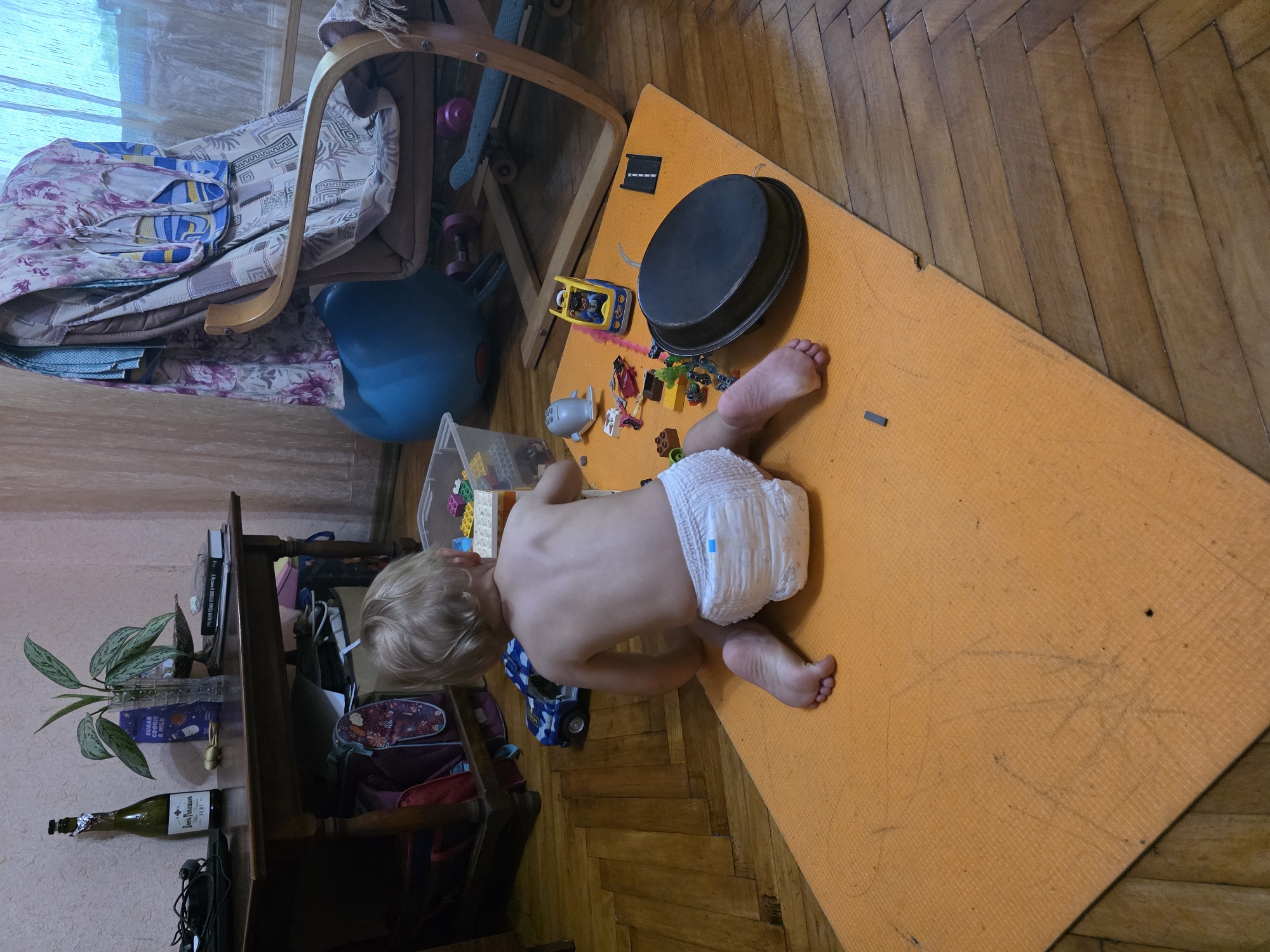 A toddler wearing a diaper sits on an orange mat on a wooden floor. Toys, including LEGO bricks, a toy car, and a small grey airplane, are scattered around the child. In the background, a wooden table holds a bottle, a plant, and books. A chair draped with fabric and a large blue exercise ball are also visible. The scene appears to be in an indoor residential setting.