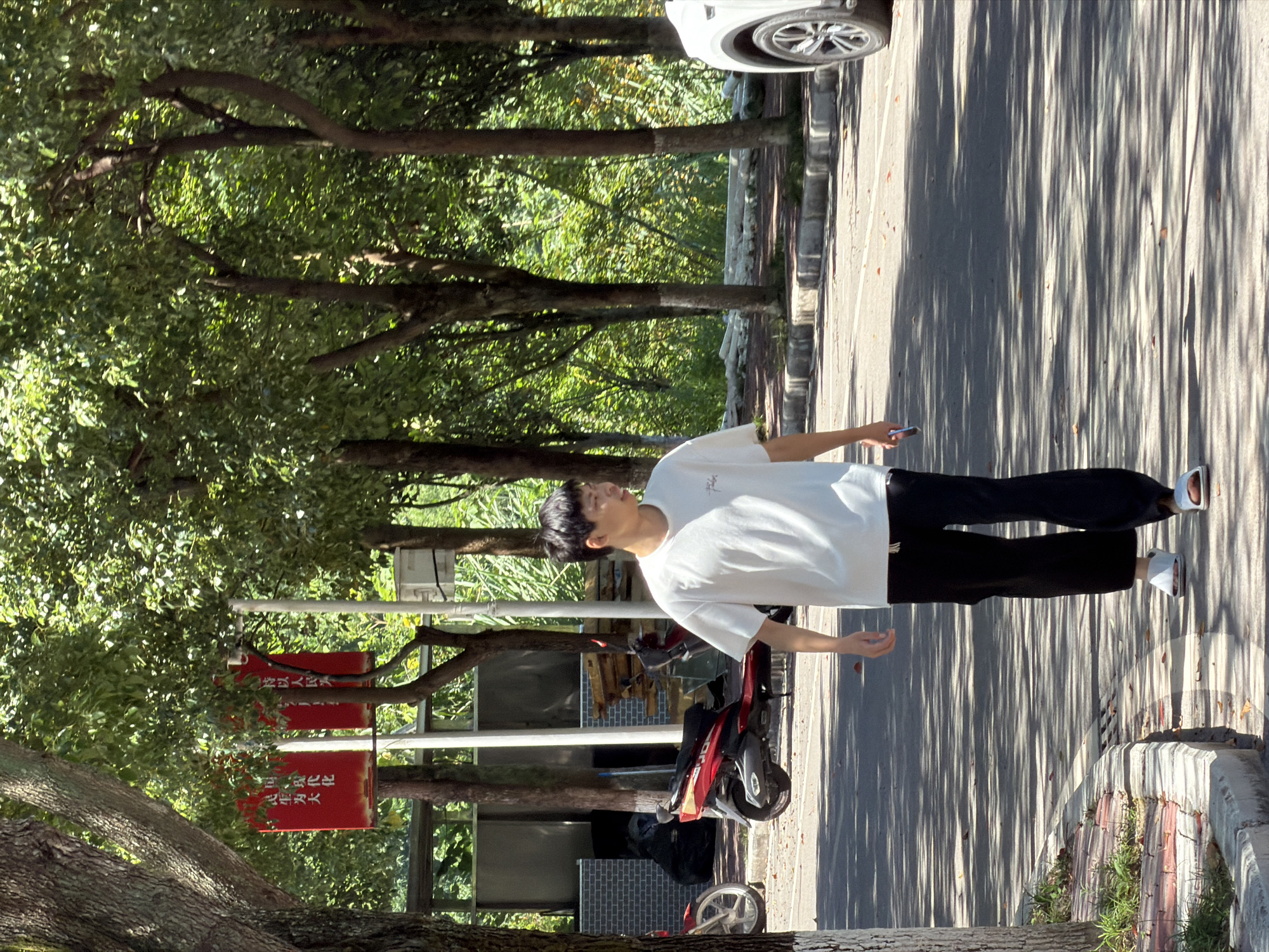 In Jiangjin, China, a young East Asian man walks along a sun-dappled road lined with trees. He is wearing a white t-shirt and loose black pants, with white slip-on sandals. In his right hand, he holds a dark-colored smartphone. To his left, a red motorcycle is parked next to a black trash bin and some stacked materials. In the background, lush green foliage and trees create a natural setting. Further down the road, the front wheel and side panel of a white SUV are visible. Red banners with Chinese characters are displayed on poles behind the motorcycle. The scene captures a casual moment on a tree-lined street.