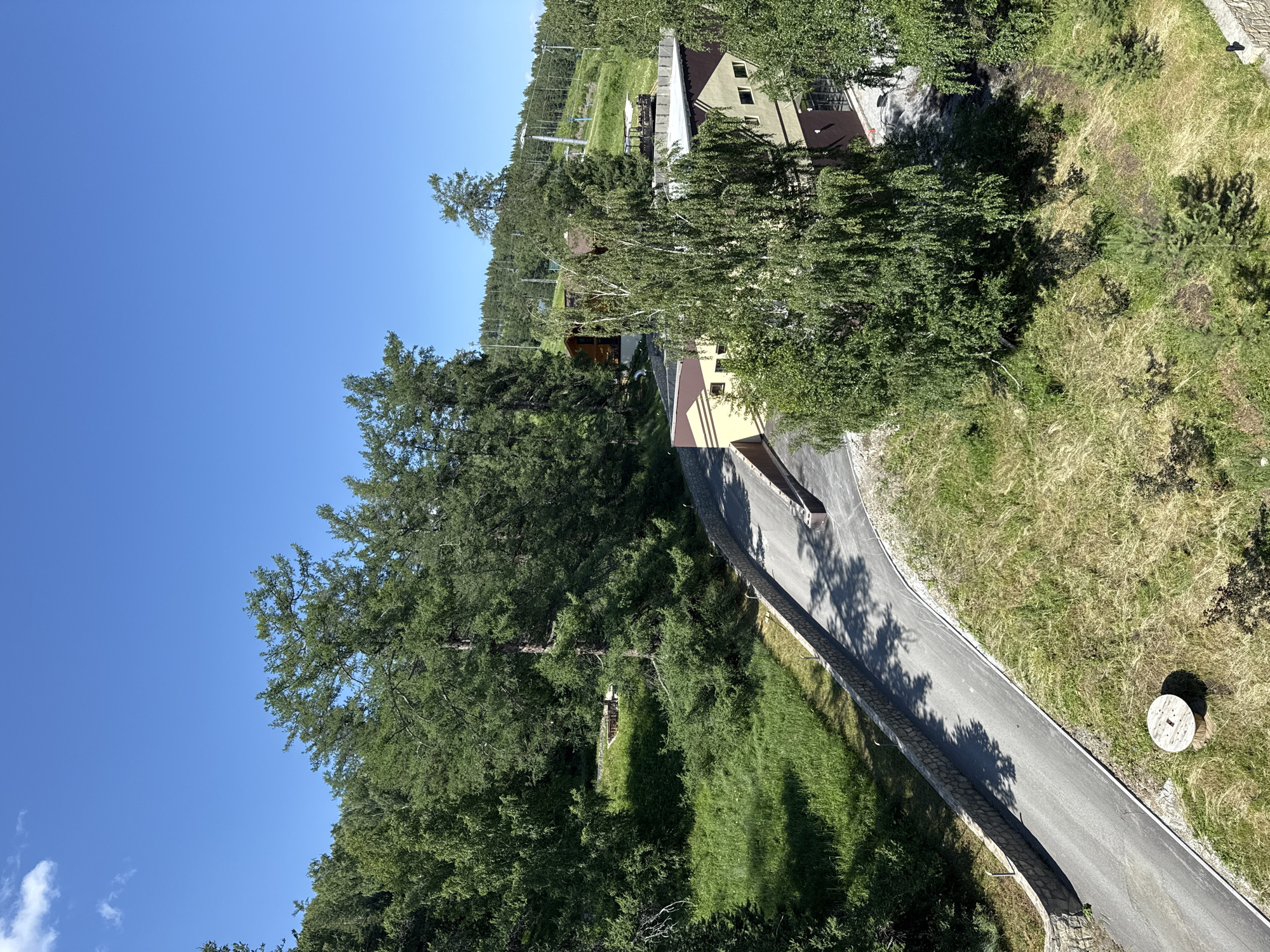The image shows a view of the Syrostan resort in Russia. In the foreground, a paved road curves uphill, bordered by a stone retaining wall on the left and grassy slopes on the right. A large, wooden spool is situated on the grassy area. A modern building with a beige and brown facade is visible on the right side of the road. In the background, a forest of tall green trees covers the landscape, with some buildings nestled among them. A ski lift and ski slopes are also discernible on a distant hill under a clear blue sky with a few scattered clouds. The overall scene depicts a tranquil, natural environment within a resort setting.