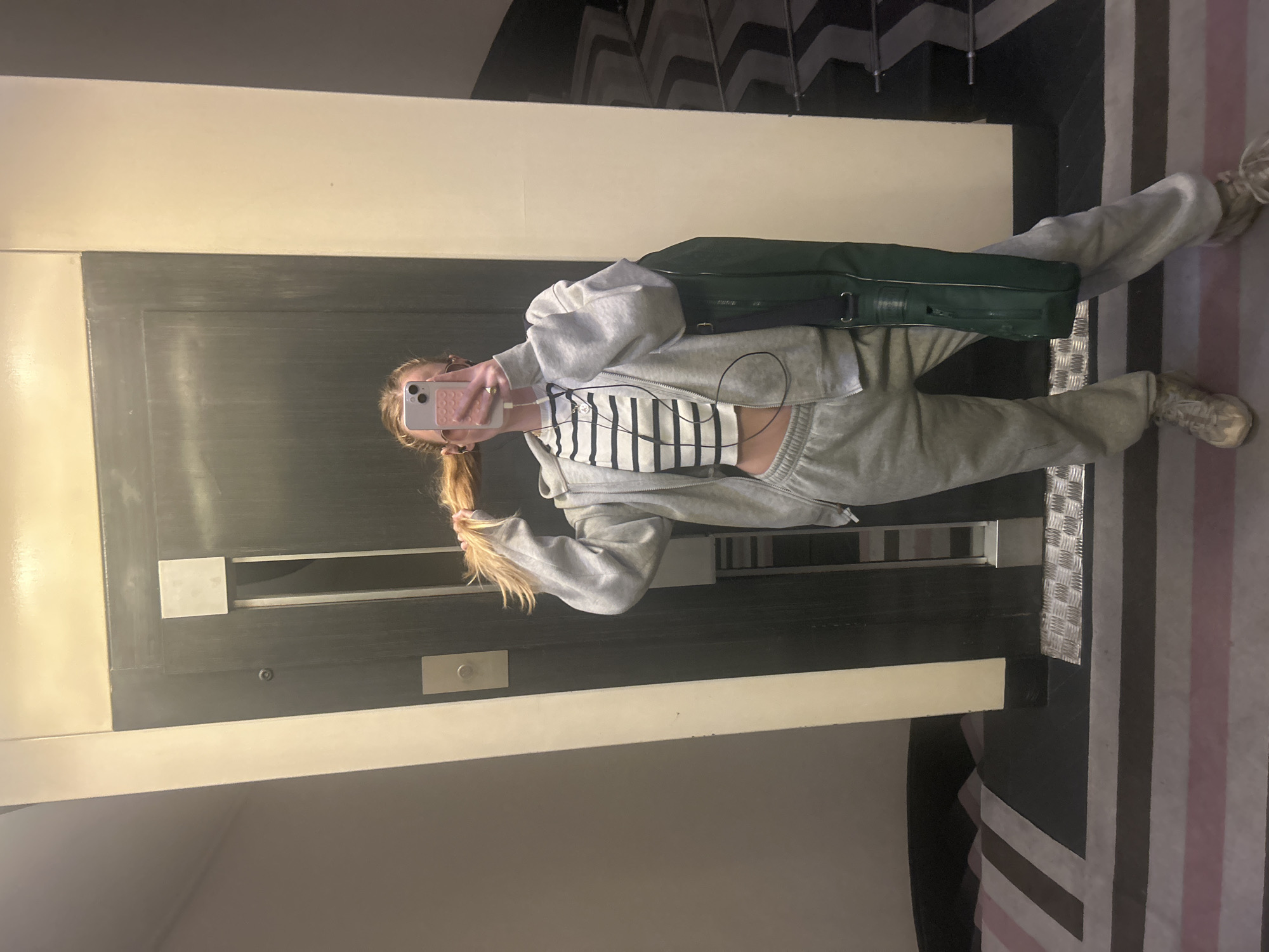 A person is taking a mirror selfie. The person has blond hair pulled back in a ponytail, and is wearing a gray sweat suit with a black and white striped top. The person is holding a pink phone. A dark green bag is slung across the person's chest. The room has a black and white patterned wall and a black and white striped floor. The person is standing in front of an elevator. The location of the scene is Unknown, Unknown.