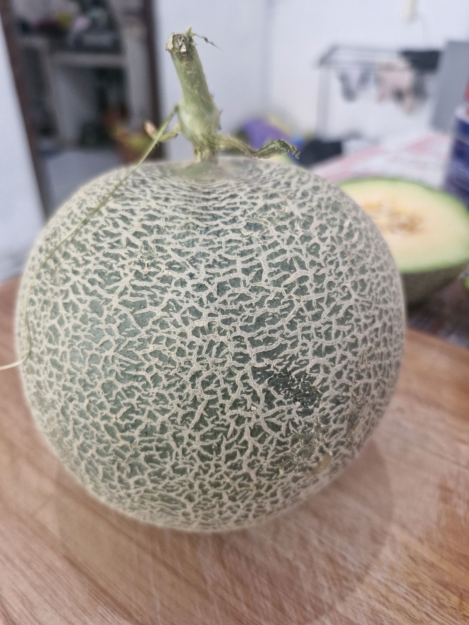 A whole cantaloupe rests on a wooden cutting board in what appears to be a kitchen. The cantaloupe is round with a green rind covered in a web-like pattern of lighter green or beige lines. A small stem protrudes from the top of the melon. To the right and out of focus, a slice of cantaloupe is visible. The background is blurred, showing what seems to be a countertop with various items and a drying rack. No individuals are present in the frame, and no specific location in Rantau, Indonesia can be identified from the visual information provided.