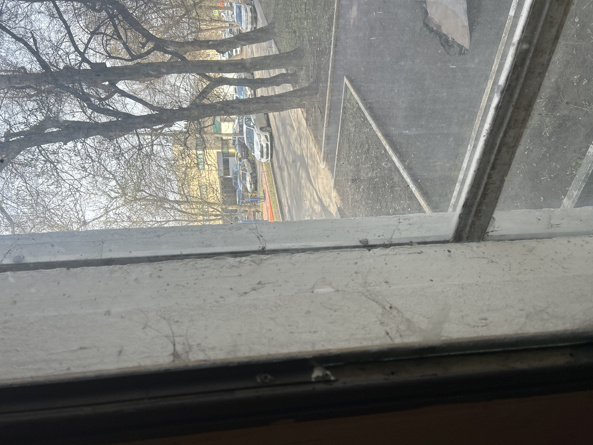 View from an interior window in Moscow, Russia. The foreground shows the window frame, dusty and aged. Through the dirty window, the exterior view includes bare trees lining a street with parked cars. Buildings are visible in the background. A patch of ground is seen directly outside the window.