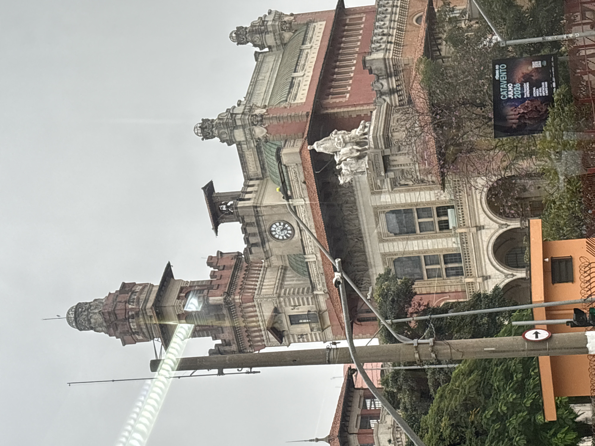 This is a photograph of the Catavento Cultural e Educacional, a science museum located in the city of São Paulo, Brazil. The building, a former Palace of Justice, exhibits a neo-classical architectural style with red brick and white stone facades. A prominent clock tower is visible on the left side of the building, and a detailed sculpture of people and horses adorns the facade. The sky is overcast, contributing to a muted lighting condition. In the foreground, a traffic light and a street sign with a white arrow are partially visible, along with some vegetation and a billboard advertising "FÉRIAS NO CATAVENTO JULHO 2026" (Catavento Holidays July 2026). The photograph appears to have been taken from a moving vehicle due to the streak of light across the upper portion of the image, likely from reflections on a window.