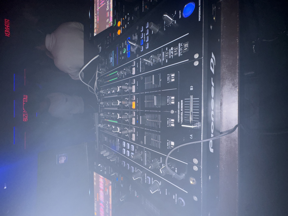 The image features a Pioneer DJ mixer, with a multitude of knobs, buttons, and sliders. Text on the mixer reads "PROFESSIONAL MIXER," "Pioneer DJ," and various DJ terminology like "CROSSFADER ASSIGN" and "MAGVEL FADER." A display screen is visible at the top of the image. Other objects and text are blurred in the background, including the word "KOYHATA," "BIZIMOW," and "04:24." It appears to be in a dimly lit environment.