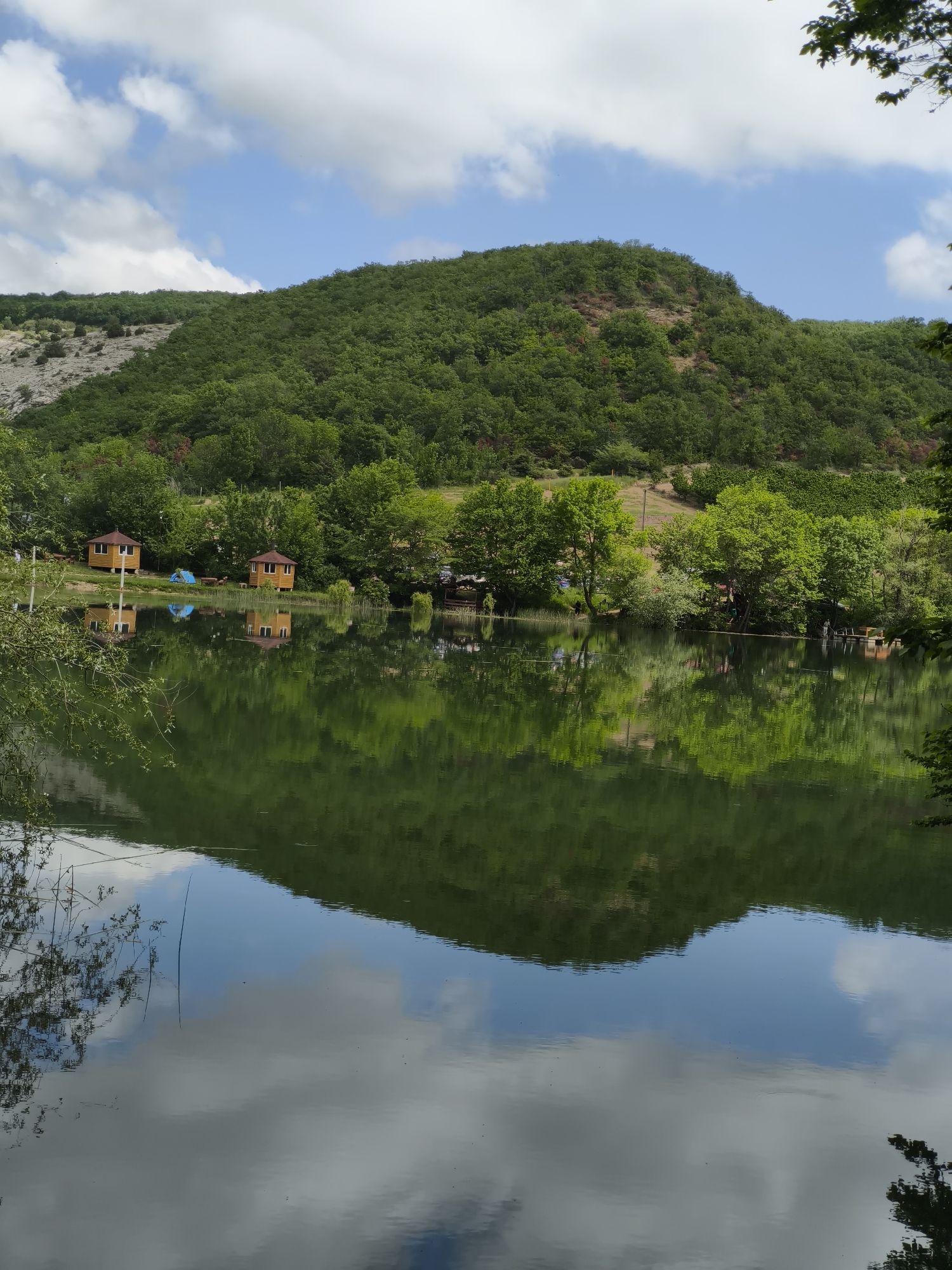 The image shows a serene lake reflecting the surrounding landscape. On the shore, there are two small wooden structures, possibly cabins or sheds. A blue tent is set up near them. Dense trees line the shore, and a grassy hill rises in the background. The sky above is partly cloudy. The reflection in the water mirrors the trees, cabins, and the hill, with additional reflections of the sky and clouds. The location is Терновский округ, Ukraine, based on the region.