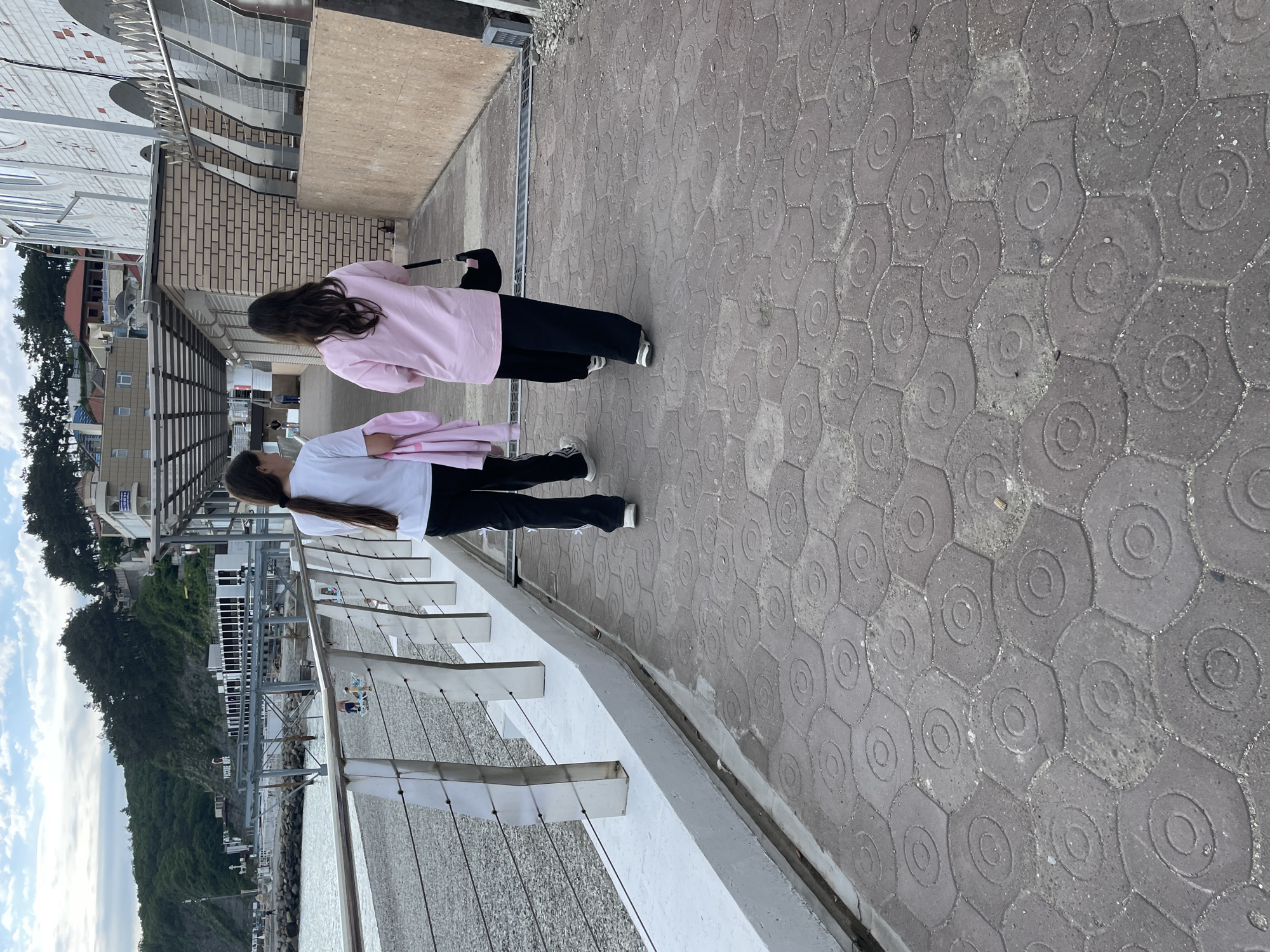 Two young women are walking along a pedestrian walkway in Olginka, Russia. One woman is wearing a white shirt and black pants, while the other is wearing a pink shirt and black pants. They are walking parallel to a railing that overlooks the coast, including the sea and forested hillside. The walkway is paved with patterned gray stones. In the background, buildings and trees can be seen on the hillside.