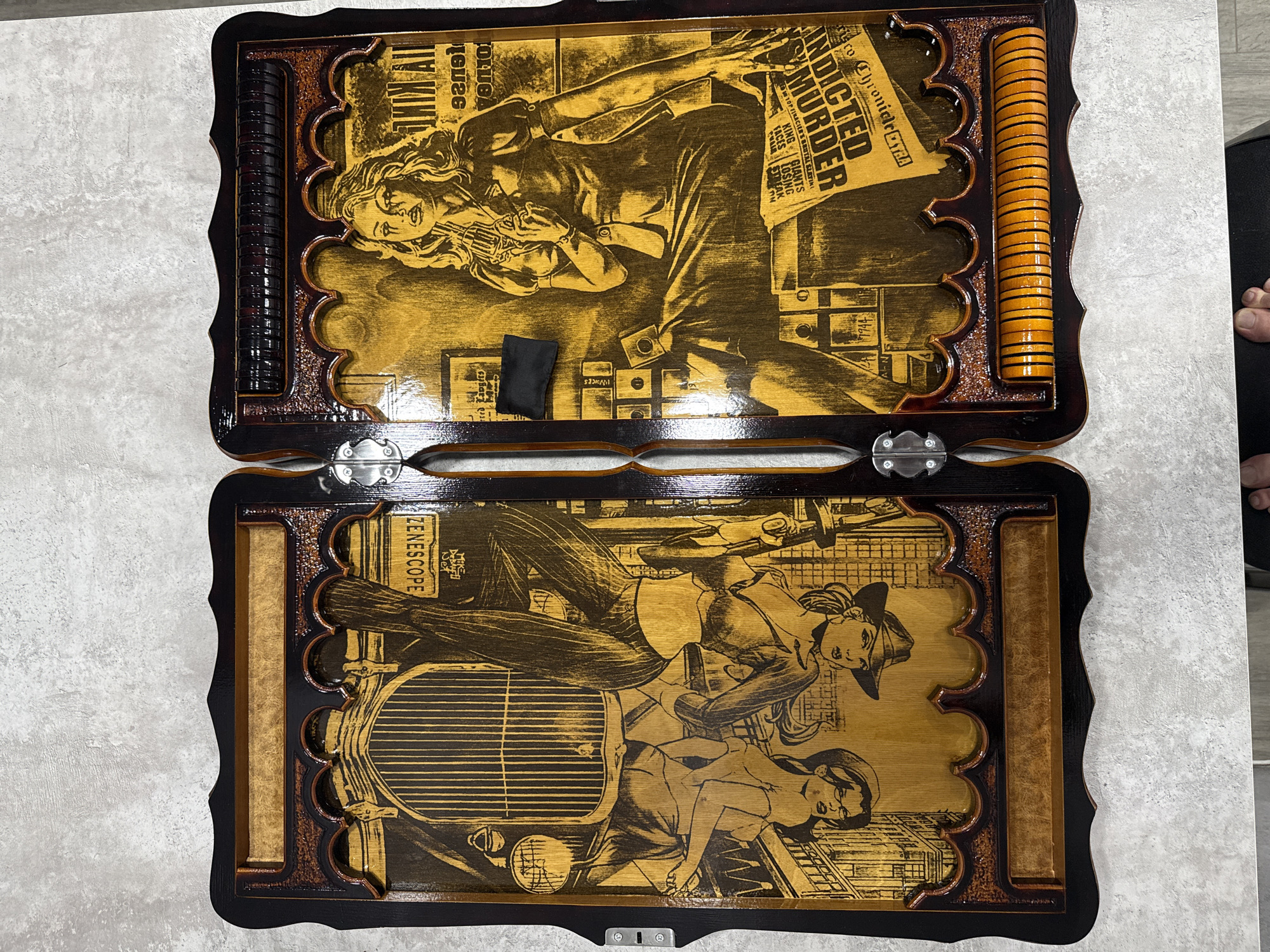 The image shows a wooden backgammon board with decorative carvings and inlays. The board is open and laid flat on a textured surface. The left side of the board features an illustration of an antique car and two figures that appear to be a couple, rendered in a dark line art style against a lighter wood background. The right side of the board displays a depiction of a woman holding a pistol, seated near a stack of what appear to be playing cards and a newspaper with the headline "INDICTED MURDER". Both illustrations have a vintage, somewhat noir aesthetic. The board has a dark, polished wood frame and a carved decorative border along the top and bottom of each side. There are also compartments at the top of each side, likely for holding game pieces, and a set of orange game pieces are visible in a holder on the bottom right. The overall impression is of a custom-made, decorative game set. There is no indication of the city of Norilsk, Russia, in the image.