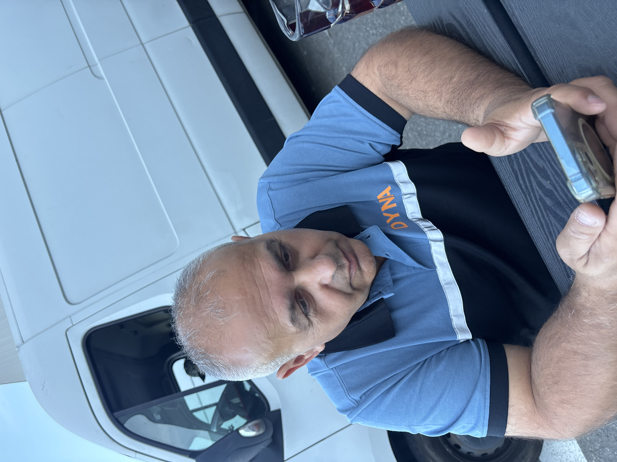 An over-the-shoulder shot shows a middle-aged man with grey hair looking down at his phone. He is wearing a blue polo shirt with black accents and orange lettering that reads "DYNA." The man is seated at a dark picnic table, and in the background, a white cargo van is parked. The scene appears to be outdoors, possibly in an industrial or commercial area, consistent with locations in the city of Boom, Belgium, known for its industrial heritage and large event venues.