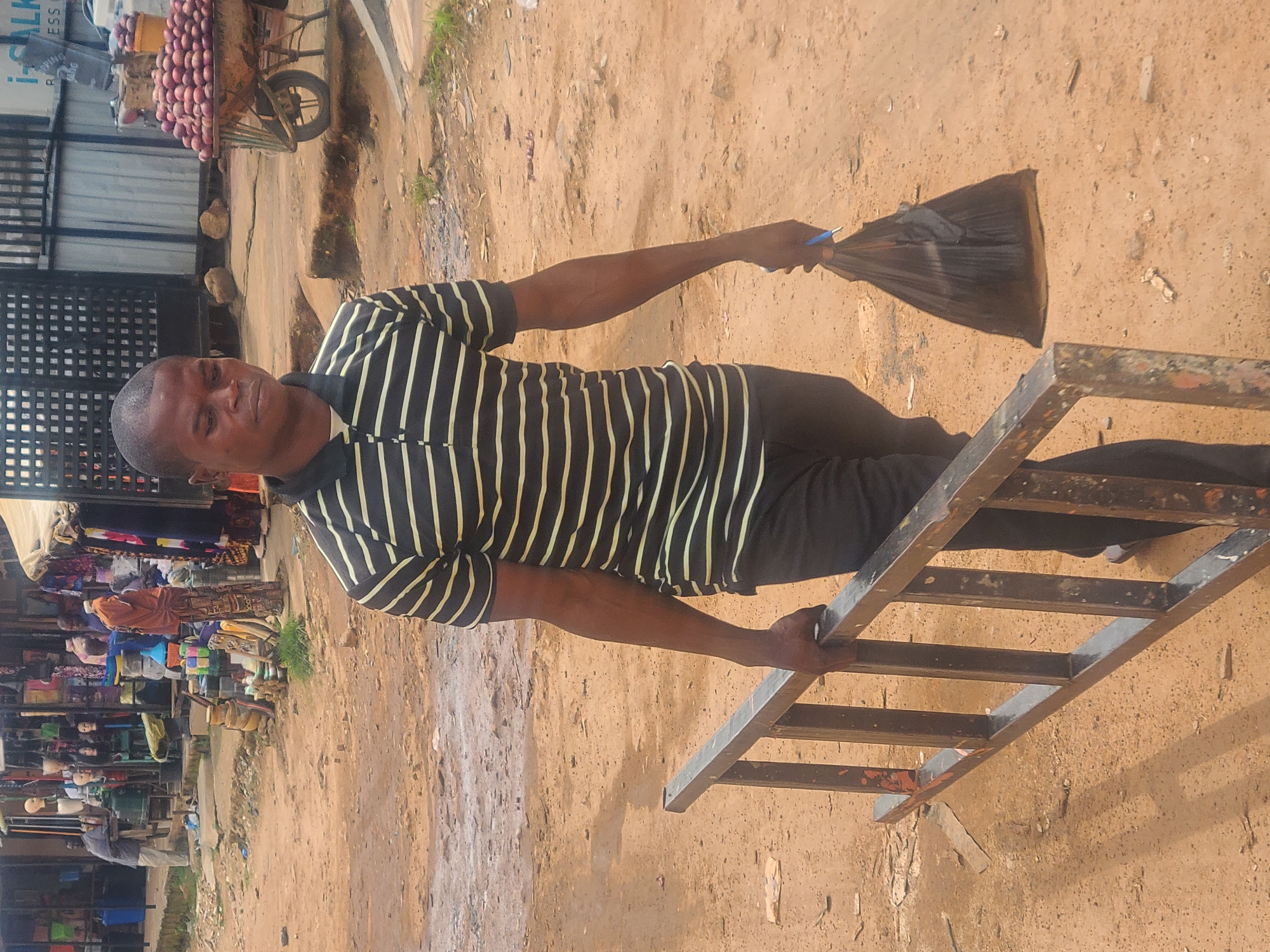 A medium shot captures a Black male in his 30s walking from right to left, carrying a metal ladder horizontally with his right hand. He is wearing a black and white horizontally striped polo shirt and dark pants. In his left hand, he holds a black plastic bag with a blue pen sticking out. The man is walking on a dirt road in a market area that appears to be in the city of Jos, Nigeria. Behind him, to his left, is a stall filled with various items, including mannequins, clothing, and baskets. To his right, there is a fruit cart overflowing with red fruits, possibly apples, and a black metal fence. The ground is covered in sand and debris, and there is some vegetation visible. The overall atmosphere suggests a busy outdoor market setting.