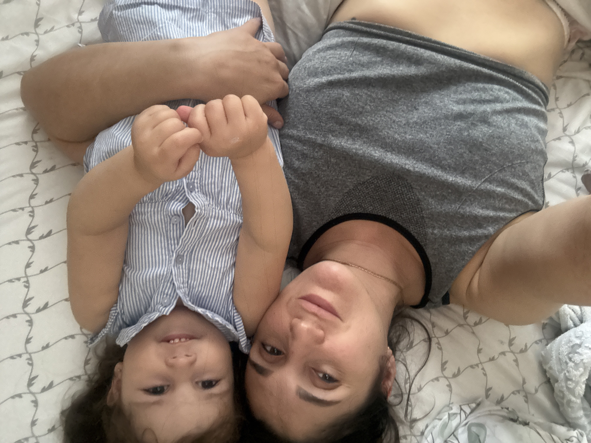 A woman and a young child lie on a bed with a white patterned comforter. The woman wears a gray tank top and a gold chain necklace. The child is dressed in a blue and white striped button-down shirt. Their hands are intertwined. The background shows a bed and bedding. It is not possible to confirm the specific location of Tobolsk, Russia from the image.