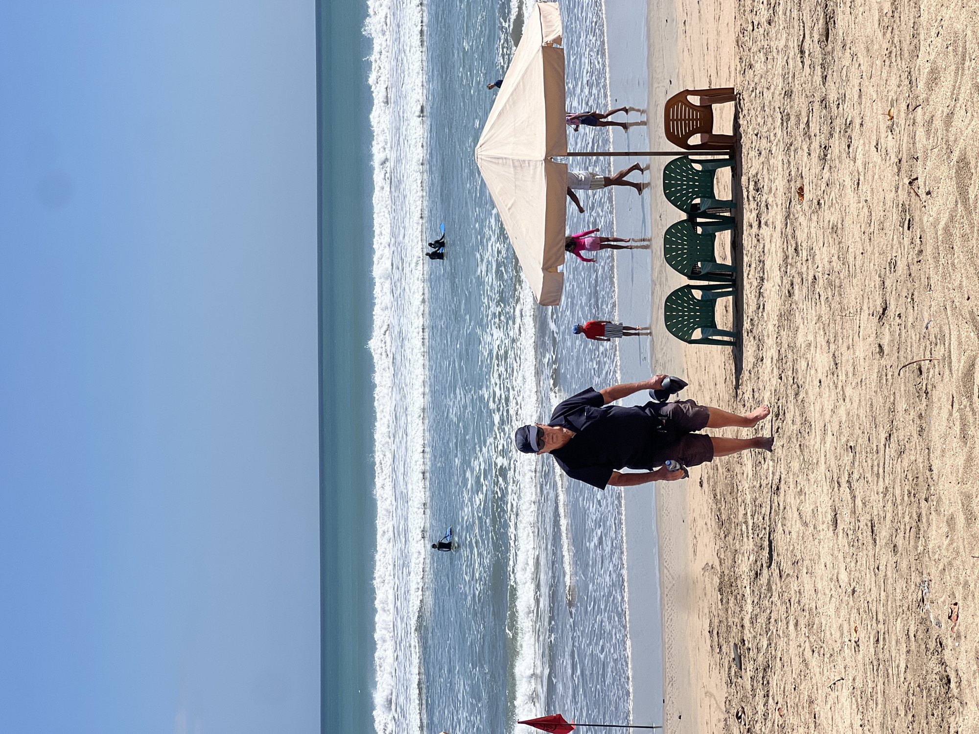 In Kuta, Indonesia, on a sunny day, a man walks along the sandy beach towards the viewer. He is wearing a dark blue short-sleeved polo shirt, dark shorts, sunglasses, and a baseball cap. He is carrying a water bottle in his left hand and what appears to be a pair of shoes in his right hand. Behind him, a red flag stands on a pole near the shoreline.
Further out in the ocean, waves crash on the shore, and several people are visible. Two individuals are in the water, one on a surfboard and another in the distance. More people, including children, are seen playing in the shallow water near the shore.
In the foreground, a large beige beach umbrella provides shade. Beneath it and to its left are four plastic beach chairs: three green ones arranged in a row, and one brown one to the right. The sand is light brown and appears to be dry in the foreground, with some debris scattered on it. The sky is clear and blue.