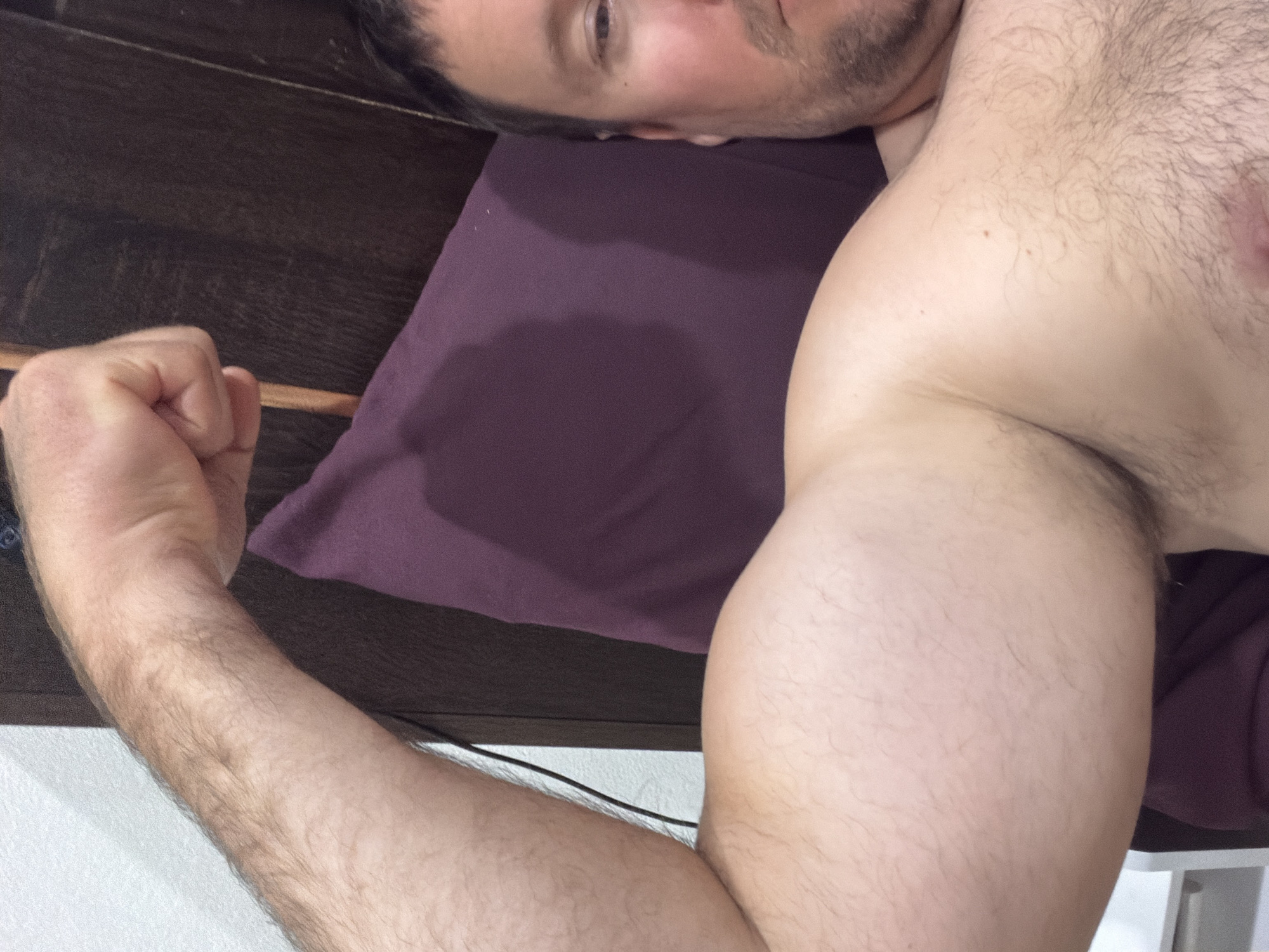 The image shows a close-up view of a man's muscular arm and chest. He is lying down, with his right arm flexed, showing his bicep. His head is partially visible on the right side of the frame, with his face turned towards the camera. He has short brown hair and a beard. He is shirtless, and his chest is covered in dark hair. He is lying on a purple pillow, and a dark wooden headboard is visible behind him. The background also shows a white wall. The man is located in a bedroom. The image does not provide any information about the city of Baan Mae Nam, Thailand, nor its specific location within that city.