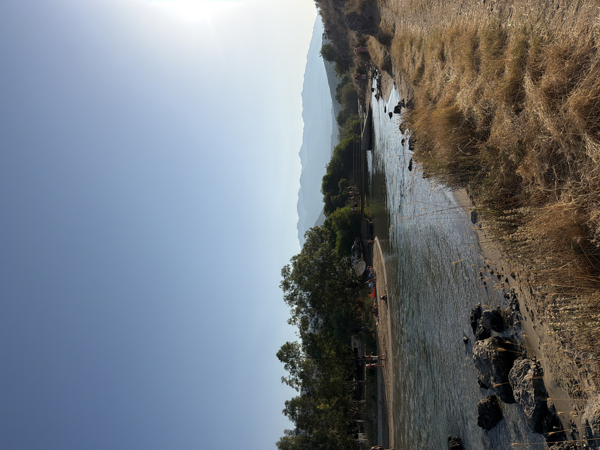 The image depicts a serene natural scene in Georgioupoli, Greece, featuring a shallow body of water, likely a river or lagoon, flowing towards a broader expanse. The water is clear, revealing rocks and pebbles beneath its surface, and a sandy shore lines its edges. Dense green trees with varying heights flank the water, casting shadows and creating a lush backdrop. In the distance, a range of hazy mountains under a clear blue sky forms a picturesque horizon.
On the sandy shore, several individuals are engaged in leisure activities. Some are standing and interacting, while others are seated or lying down, enjoying the sun. A few people appear to be wading in the shallow water. The presence of a bridge in the mid-ground suggests a pathway or crossing over the water. The overall atmosphere is one of tranquility and natural beauty, characteristic of a coastal or riverside resort area. The sun is visible in the upper right corner, indicating it is either early morning or late afternoon, casting a warm glow on the landscape. The dry, golden grass on the right side of the image adds to the rustic charm of the scene.