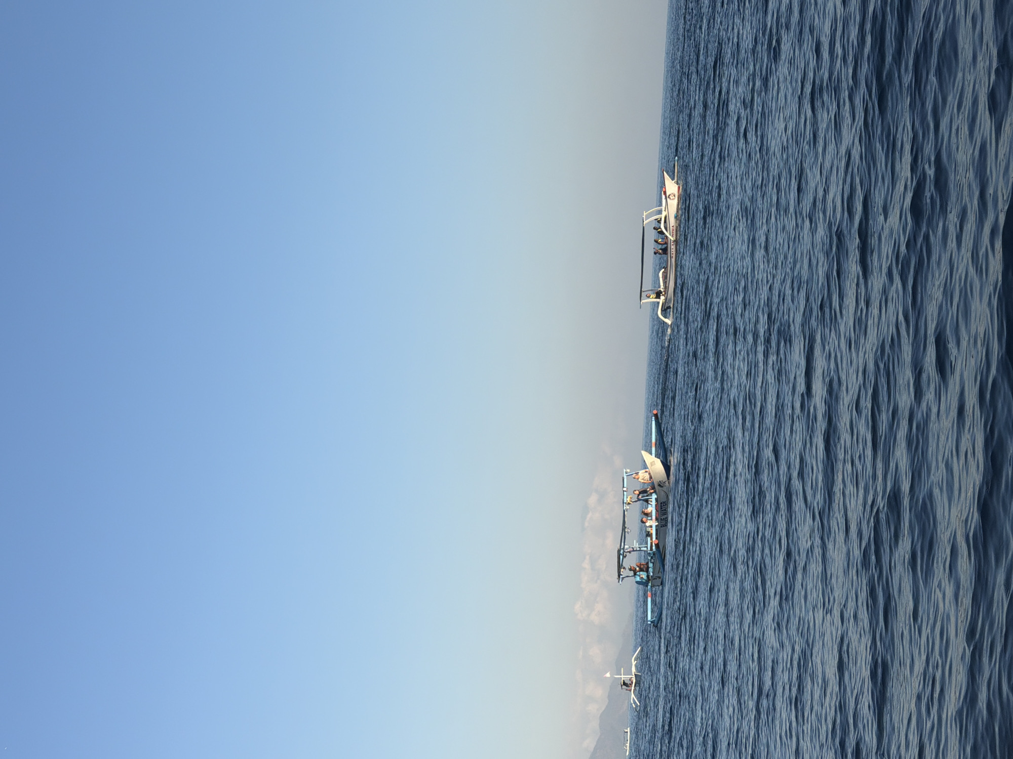 In the ocean, three boats are visible. The closest boat is a narrow, outrigger boat with a blue and white hull and a canopy. It carries several passengers, some of whom appear to be tourists. The boat is labeled "BLUE WATER". Further in the distance, another similar boat with a canopy is visible, also carrying passengers. On the far left, a smaller outrigger boat with a white flag flies in the distance. The water is choppy with small waves. In the background, a hazy coastline with mountains is visible under a clear blue sky. The overall scene depicts a day at sea, likely for recreational purposes. The image appears to be taken from a boat moving through the water.