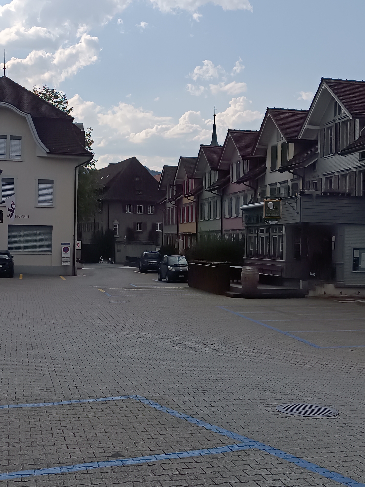 The image depicts a street scene in Appenzell, Switzerland, characterized by traditional Swiss architecture. The foreground features a cobblestone street with designated parking spaces marked by blue lines. Several cars are parked along the street. The buildings lining the street are multi-story, with pitched roofs and colorful facades, typical of Appenzell's historic town center. A church spire is visible in the background, rising above the rooftops. The sky is partly cloudy, suggesting fair weather. The overall impression is one of a well-maintained, picturesque European town.