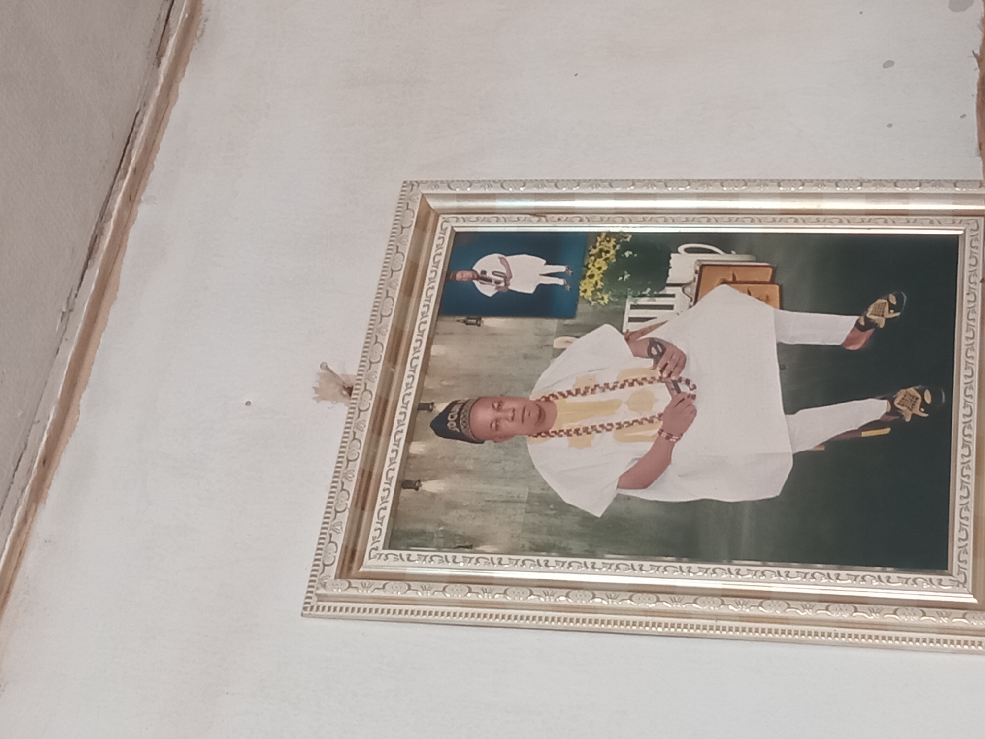 The image displays a framed portrait hanging on a plain, light-colored wall. The main subject of the portrait is a fair-skinned man, appearing middle-aged, wearing traditional Nigerian attire. He is seated, facing forward with a neutral expression. His outfit consists of a white flowing top, likely an agbada, with elaborate gold and yellow embroidery around the neck and chest, and white trousers. He wears a dark blue cap with some white, partially illegible text, possibly "ADOH". A multi-strand gold-colored beaded necklace and matching bracelets adorn him. His feet are visible, wearing black and gold open-toed sandals.
The backdrop of the portrait features a deep green and blue-green gradient, with some light-colored objects on the right that resemble stacked books or boxes, and a white pitcher or jug. To the left, a smaller, faded image of another person, also in white attire, is subtly incorporated into the background. Some yellow floral elements are visible behind the man on the right side. The portrait is enclosed in an ornate, possibly gilded, frame with intricate patterns and carvings.
The wall on which the portrait hangs is off-white or cream, showing some textural imperfections. In the upper left corner of the original image (which would be the upper right if rotated correctly), a wall corner with minor damage or a gap is visible. A small, decorative, tassel-like object is suspended near the top right corner of the frame. The scene captures a formal portrait, likely taken in a studio setting, emphasizing the subject's cultural attire and dignified pose. The context of Shagamu, Nigeria, aligns with the traditional attire depicted.