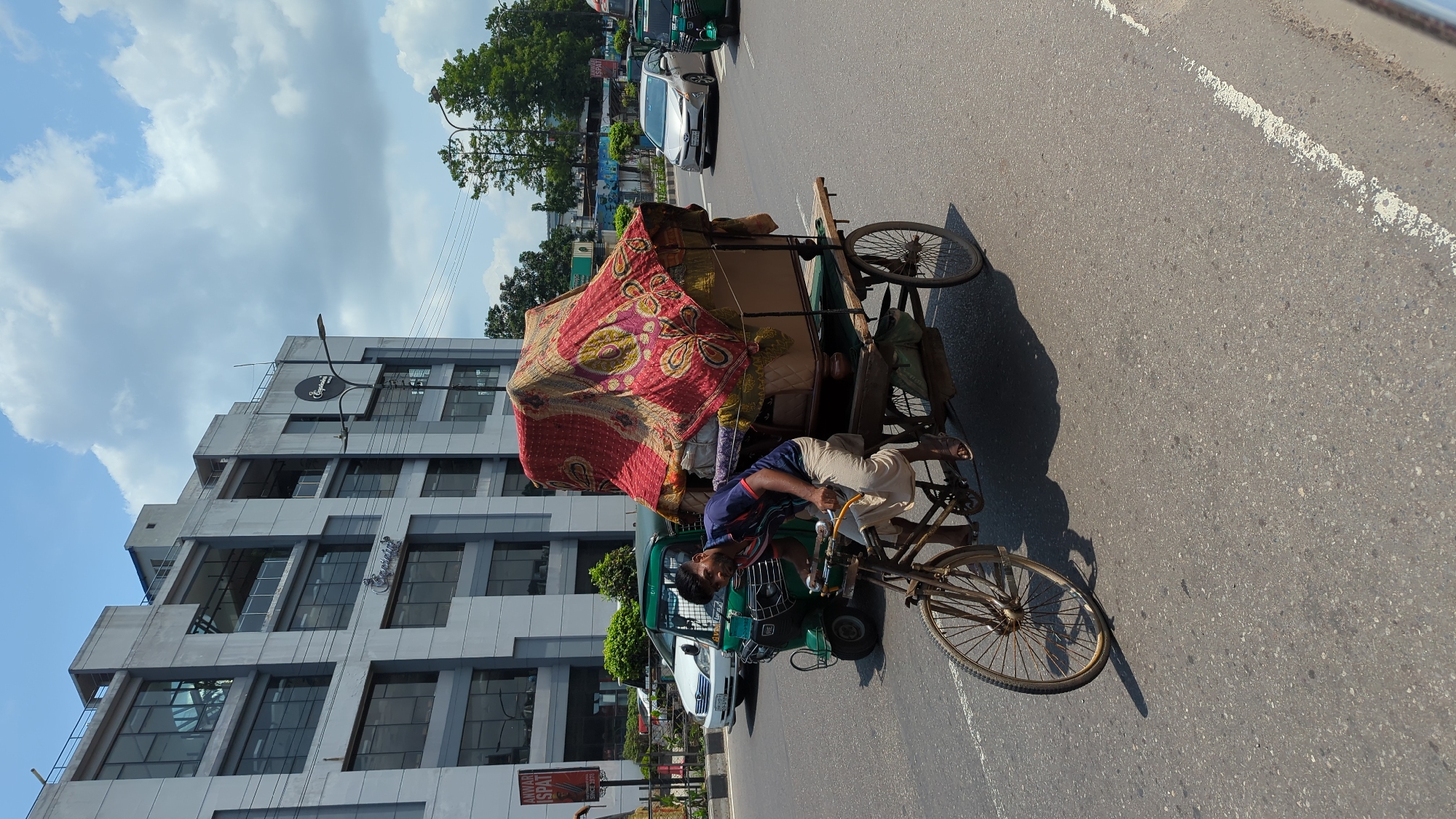 A man pedals a bicycle rickshaw piled high with goods covered by a red patterned cloth on a city street. He wears a blue shirt and beige pants. A green CNG auto rickshaw follows behind. Cars and other vehicles are in the background. The street is lined with trees and buildings. A multi-story modern building with a sign on its facade is visible. The sky is partly cloudy. The location appears to be in Dhaka, Bangladesh.