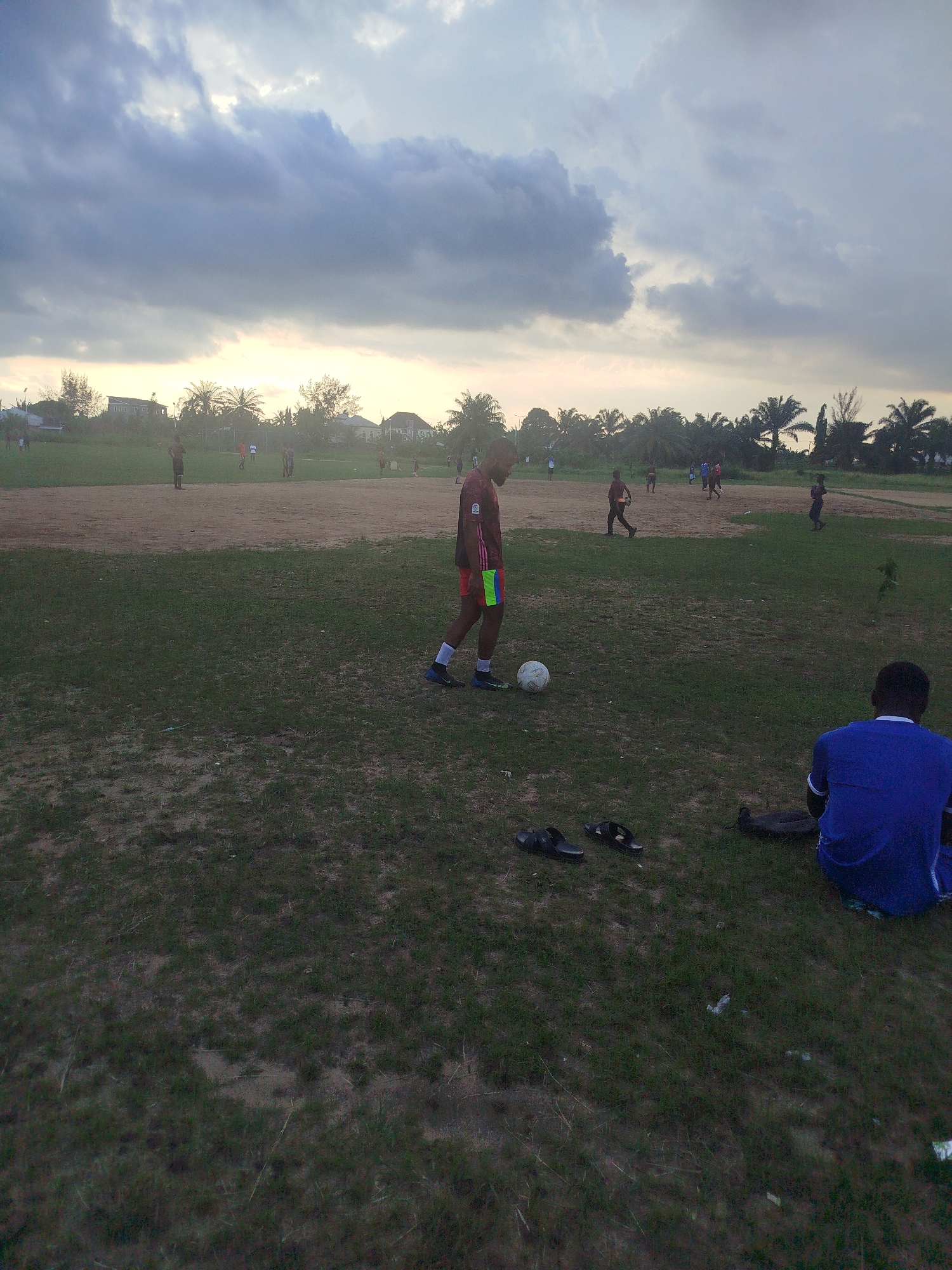 The image shows a soccer field in Ikot Mbonde, Nigeria under an overcast sky. Several individuals are scattered across the field; some appear to be engaged in a soccer game. In the foreground, a male stands near a soccer ball wearing a red and black shirt with red shorts. Nearby, a person wearing a blue shirt is seated on the ground. A pair of sandals rests on the grass between them. The field has patches of grass and bare earth. In the background, buildings and palm trees are visible.