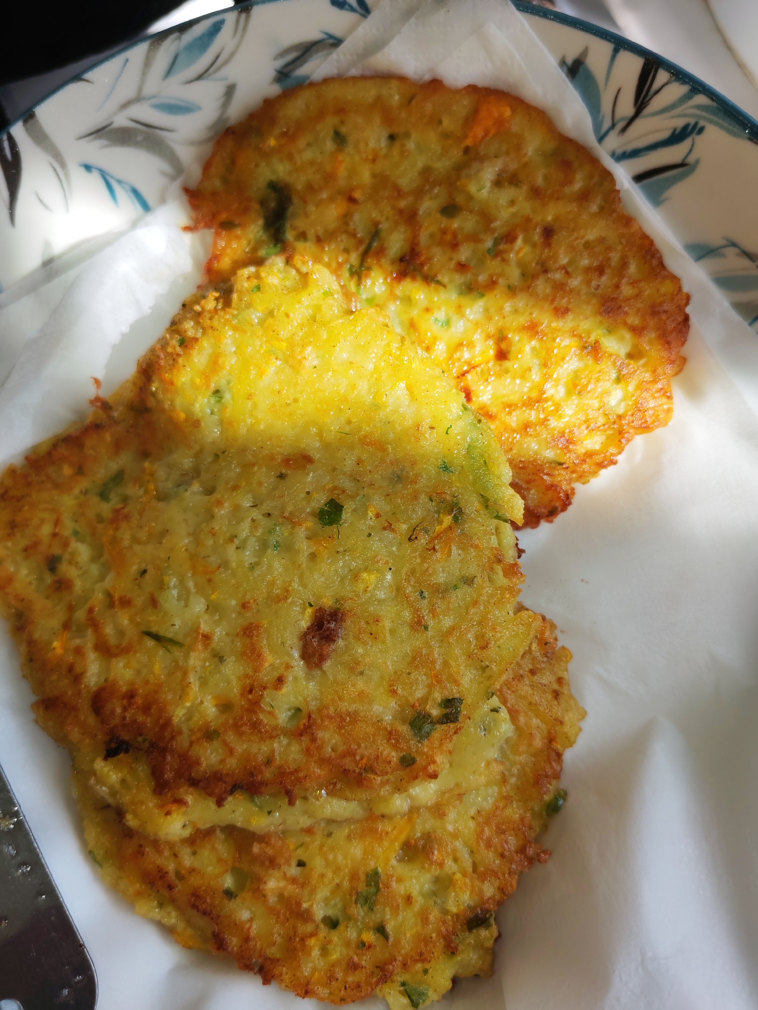 A close-up, overhead shot shows three golden-brown, flat vegetable fritters stacked on a white paper liner. The fritters have a slightly irregular, circular shape and appear to be made from grated vegetables, possibly potatoes and carrots, mixed with herbs. The edges of the fritters are crisp and slightly darker than the centers, indicating they have been fried. A portion of a decorative white plate with blue leaf patterns is visible in the upper right corner. The lighting suggests it is daytime, with a bright light source creating a glare on the top fritter. There is no information in the image to determine its location in Izhevsk, Russia.