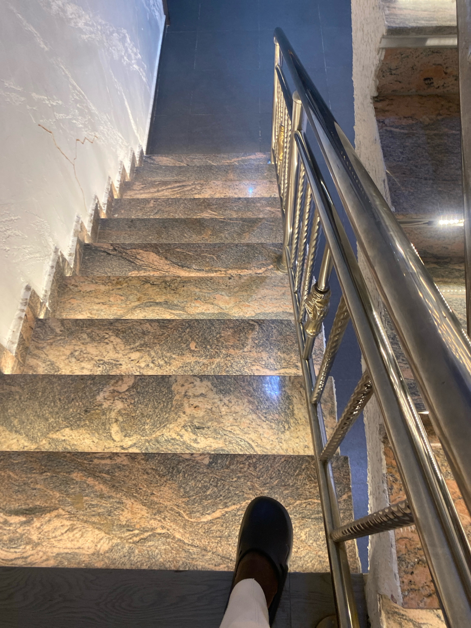 A high-angle, full shot captures a person's foot wearing a black slip-on shoe descending a modern, well-lit marble staircase. The staircase features polished granite steps with swirls of pink, brown, and gray, illuminated by recessed lighting embedded in the wall. A sleek, silver metal handrail with decorative vertical spindles runs along the right side of the stairs. The wall to the left is textured white, while the floor at the top of the stairs appears to be dark tile. The scene is devoid of any identifying landmarks that would indicate the specific city of Barden Yana, Nigeria.