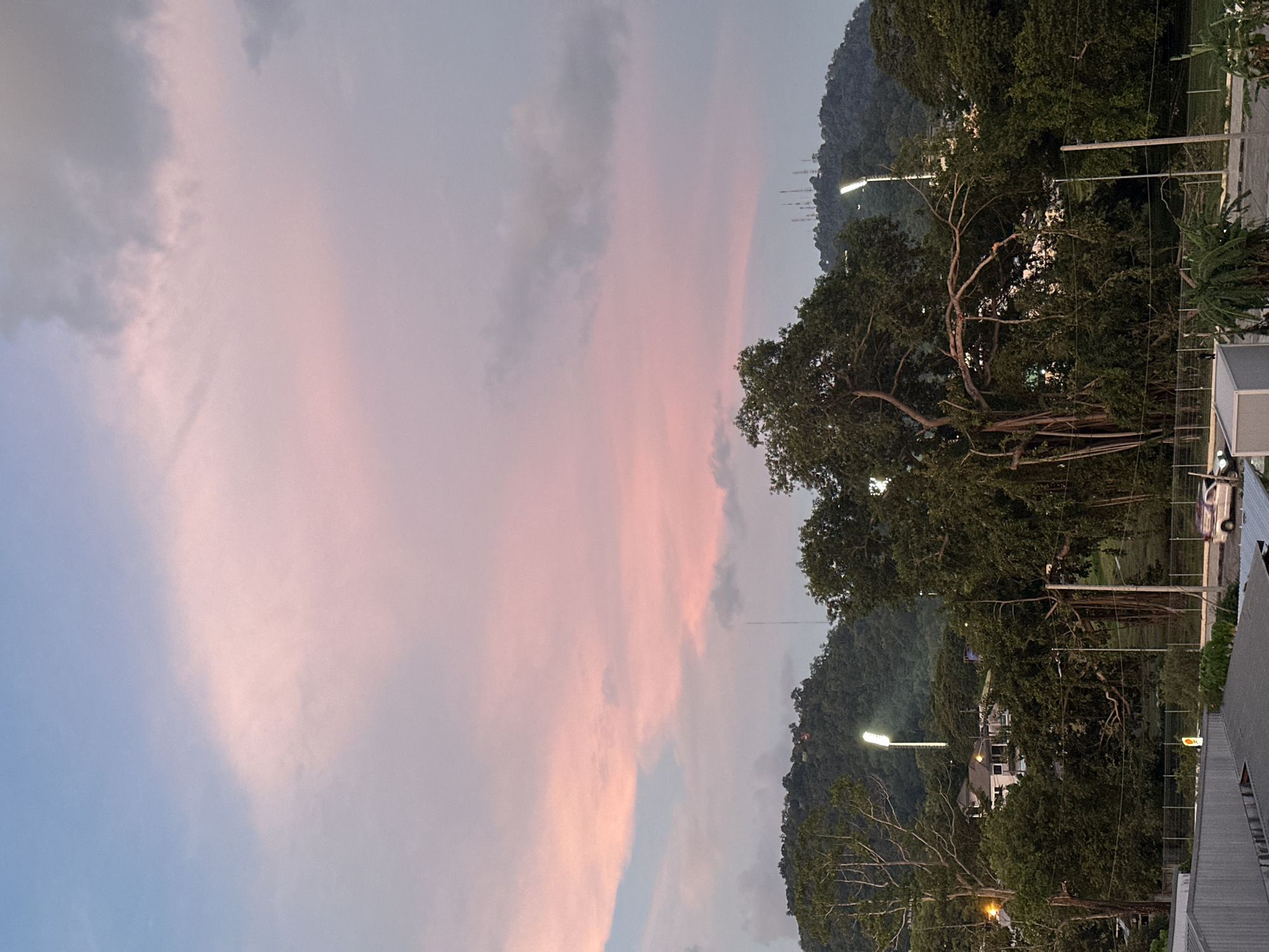 The image depicts a twilight scene overlooking a city with lush green hills in the background. The sky is painted with soft hues of pink and blue from the setting sun, with scattered clouds. In the mid-ground, there are dense trees and some buildings, including what appears to be a residential area with white houses. Stadium lights are visible on the hills, illuminating the landscape. The foreground shows the rooftops of some structures, a street with a parked white car, and fencing. The overall impression is of a peaceful evening in an urban area adjacent to natural, hilly terrain. The location is identified as Wichit, Thailand.