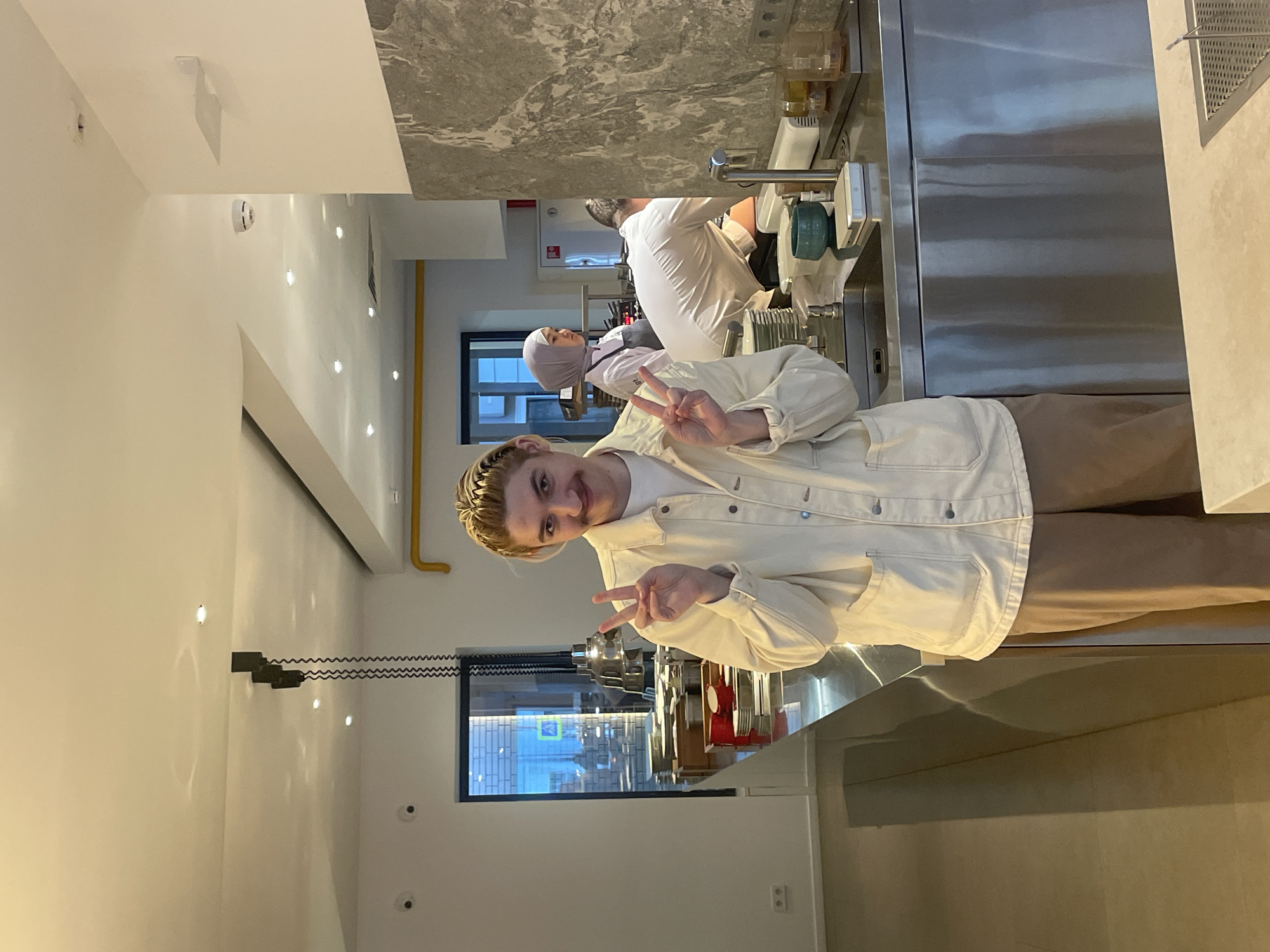 A young person is standing in what appears to be a restaurant kitchen. They are wearing tan pants, a white t-shirt, and a white denim jacket, and they are holding up two fingers in a peace sign with both hands. In the background, two chefs wearing white uniforms and head coverings are working. The kitchen has stainless steel countertops and equipment. A window is visible, reflecting the outside environment, possibly indicating an urban setting. Based on available resources, it is impossible to definitively confirm the location in the city of Moscow, Russia, without further information.