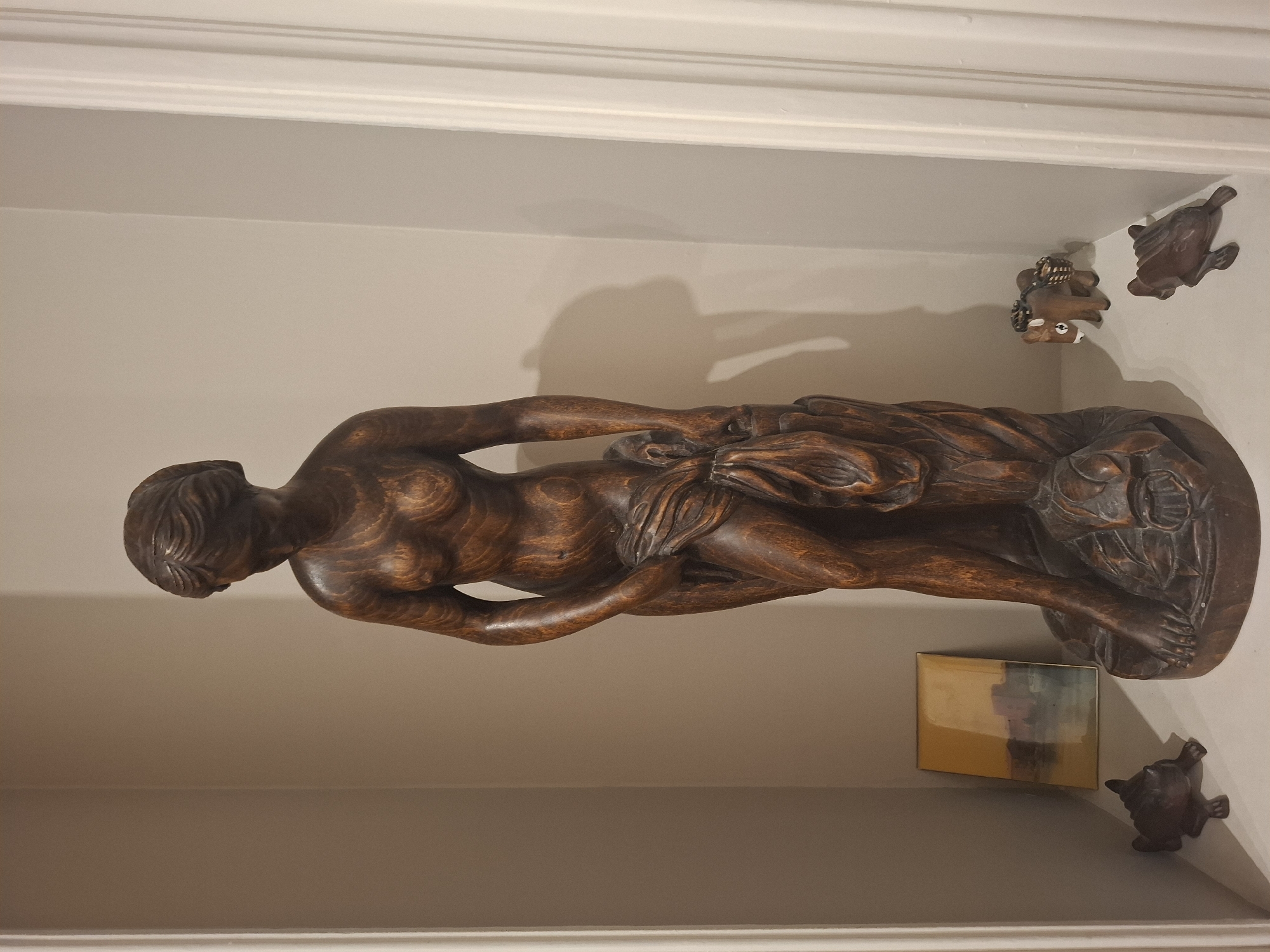 This is a full shot of a wooden statue of a nude woman in a recessed shelf. The statue is dark brown and intricately carved, depicting the woman with her head bowed and draped in fabric. The shelf is painted a neutral light color. To the left of the statue, there is a small framed painting with abstract hues of yellow and blue, and two small wooden figurines that appear to be toads. To the right of the statue, there are two more small wooden figurines, one resembling a donkey with baskets on its back and another toad. The scene is well-lit, casting shadows on the wall behind the statue. This image was taken in Vincennes, France, within a residential setting, likely a home.