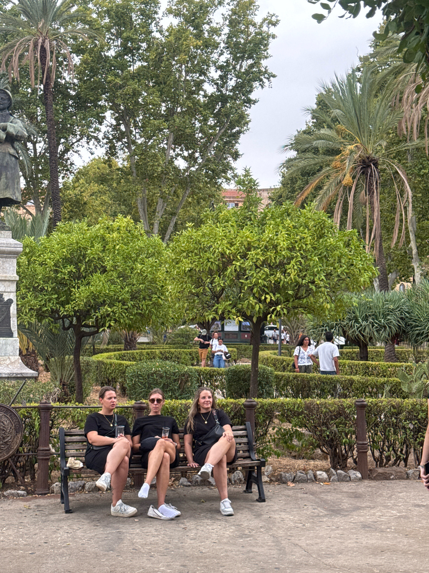 In a park in Palermo, Italy, a group of people are enjoying a day outdoors. Three young women in black dresses are sitting on a wooden bench, chatting and holding drinks. Behind them, a meticulously manicured hedge maze suggests a formal garden setting. In the background, other park-goers stroll amongst palm trees and lush greenery. A bronze statue stands to the left, hinting at the historical significance of the location. The scene is bathed in soft, diffused light under an overcast sky.
