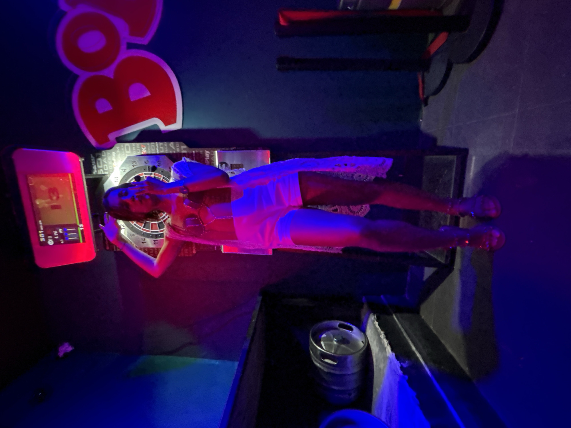A person is centrally positioned in a dimly lit recreational space, likely in Calella, Spain, illuminated by neon blue and magenta lighting. The individual, appearing female, wears white shorts, a light top, and a long, sheer white lace-patterned cover-up, with sandals visible on their feet. One hand is raised towards their mouth/face, and the other arm is slightly elevated, poised near an electronic dartboard machine directly behind them. The dartboard's digital display shows "501" and the number "13". To the left of the person, a dark shelf holds a silver metal beer keg. To the right, a large red letter "B" with a bubbly font is affixed to a dark wall, with two dark vertical poles visible on the floor below it. The floor is dark and reflective, showing hints of the colored lighting.